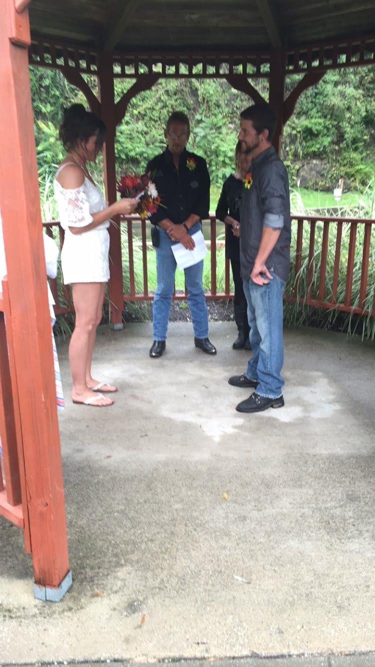 A couple exchanges vows while standing in a gazebo surrounded by greenery on a gray day.
