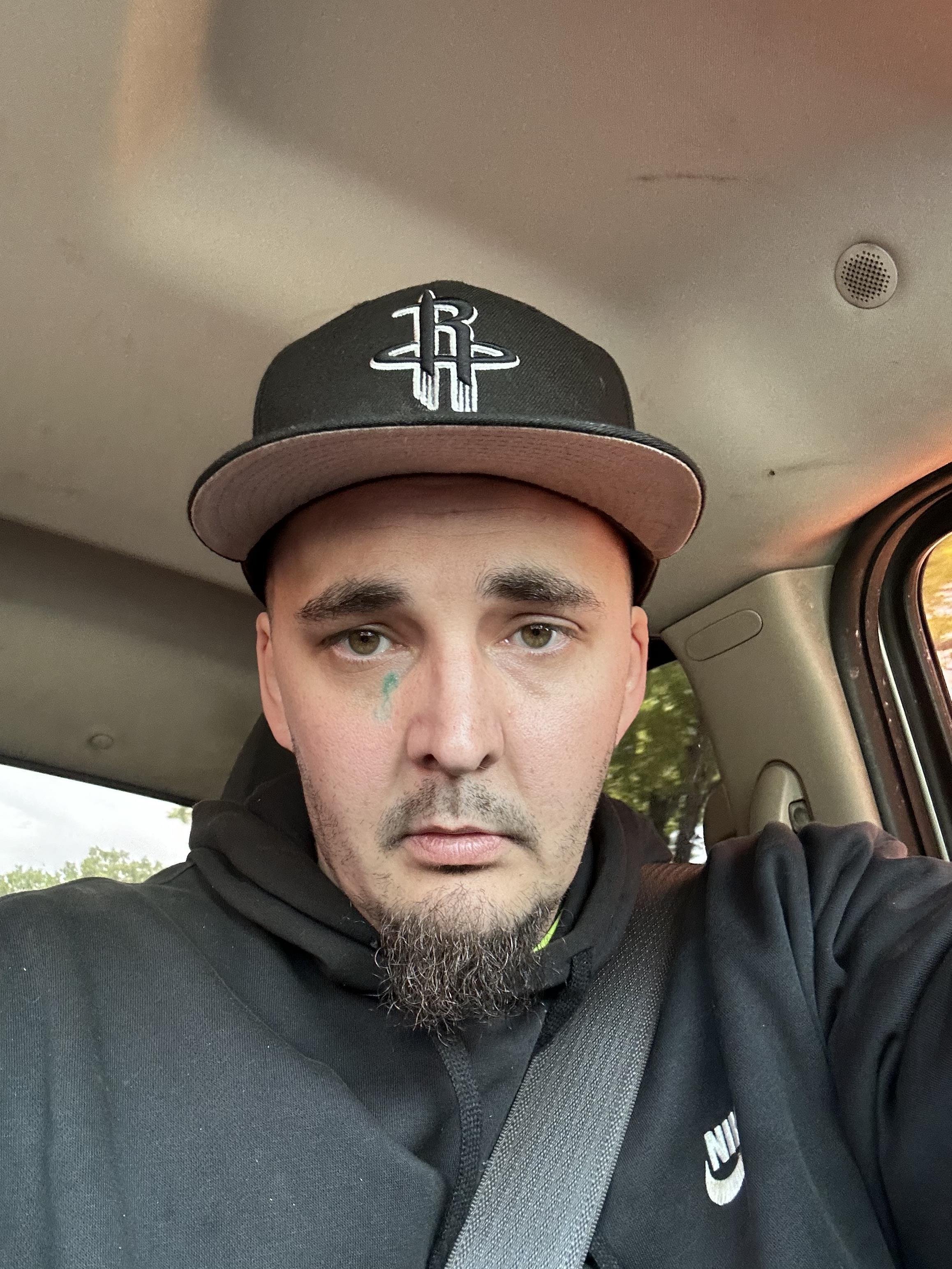 A man in a black hoodie and cap looks directly at the camera while sitting in a vehicle.