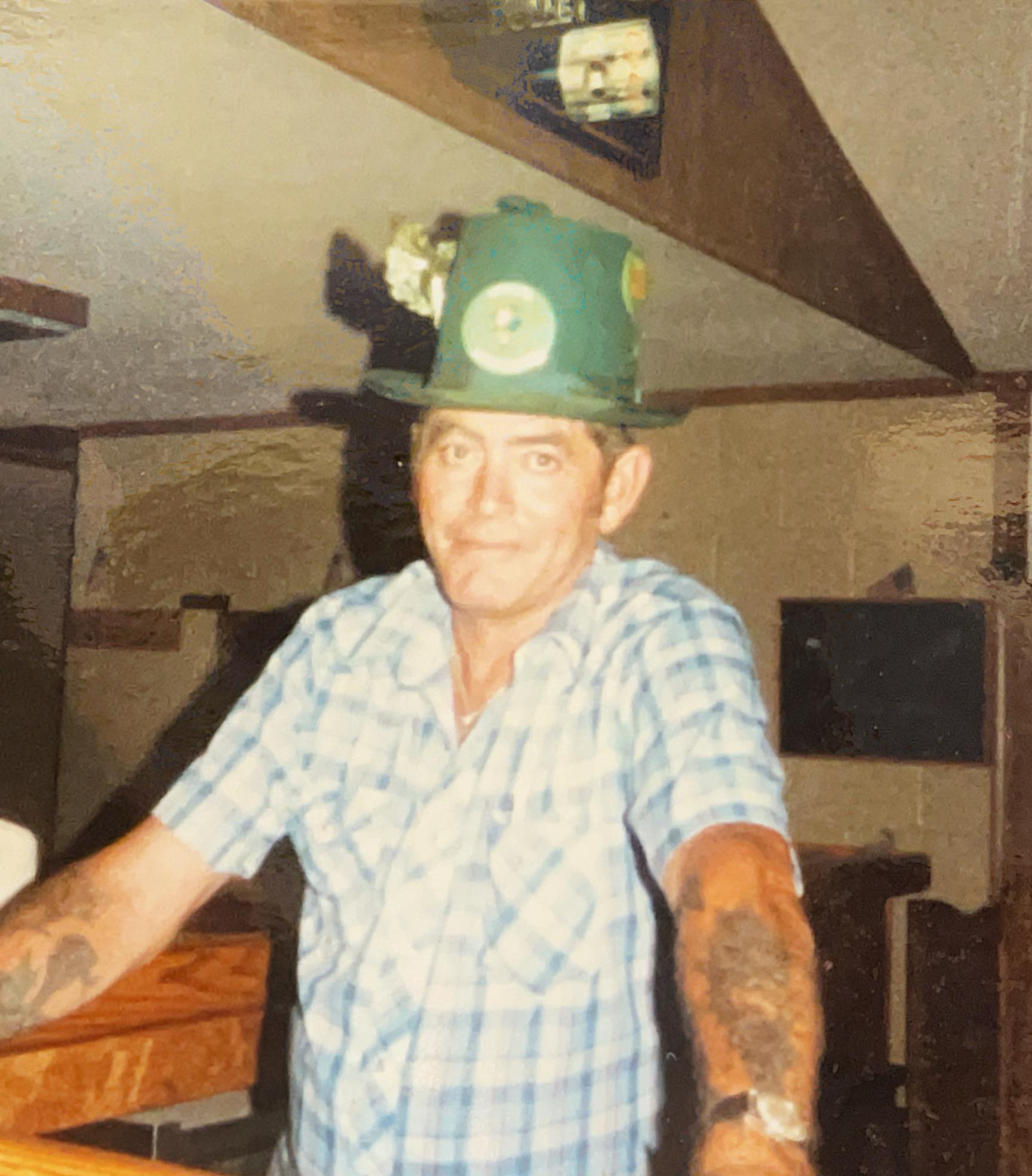 A man with tattoos leans on a bar wearing a playful green hat with a flower.