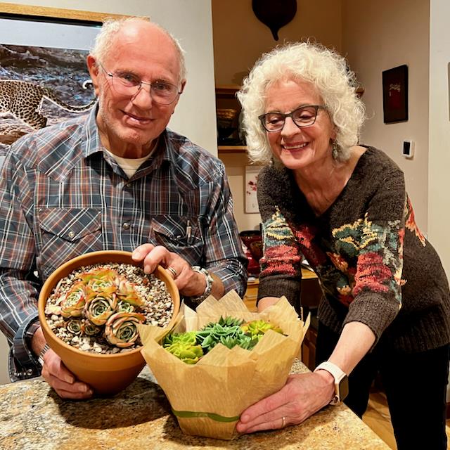 Two seniors smile as they hold beautiful succulent arrangements at home in the evening.