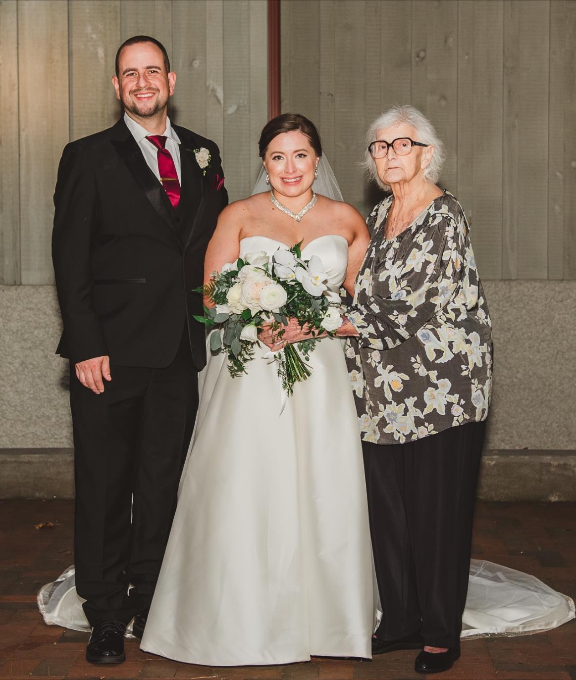 A joyful bride stands with her husband and grandmother, celebrating love and family at dusk.