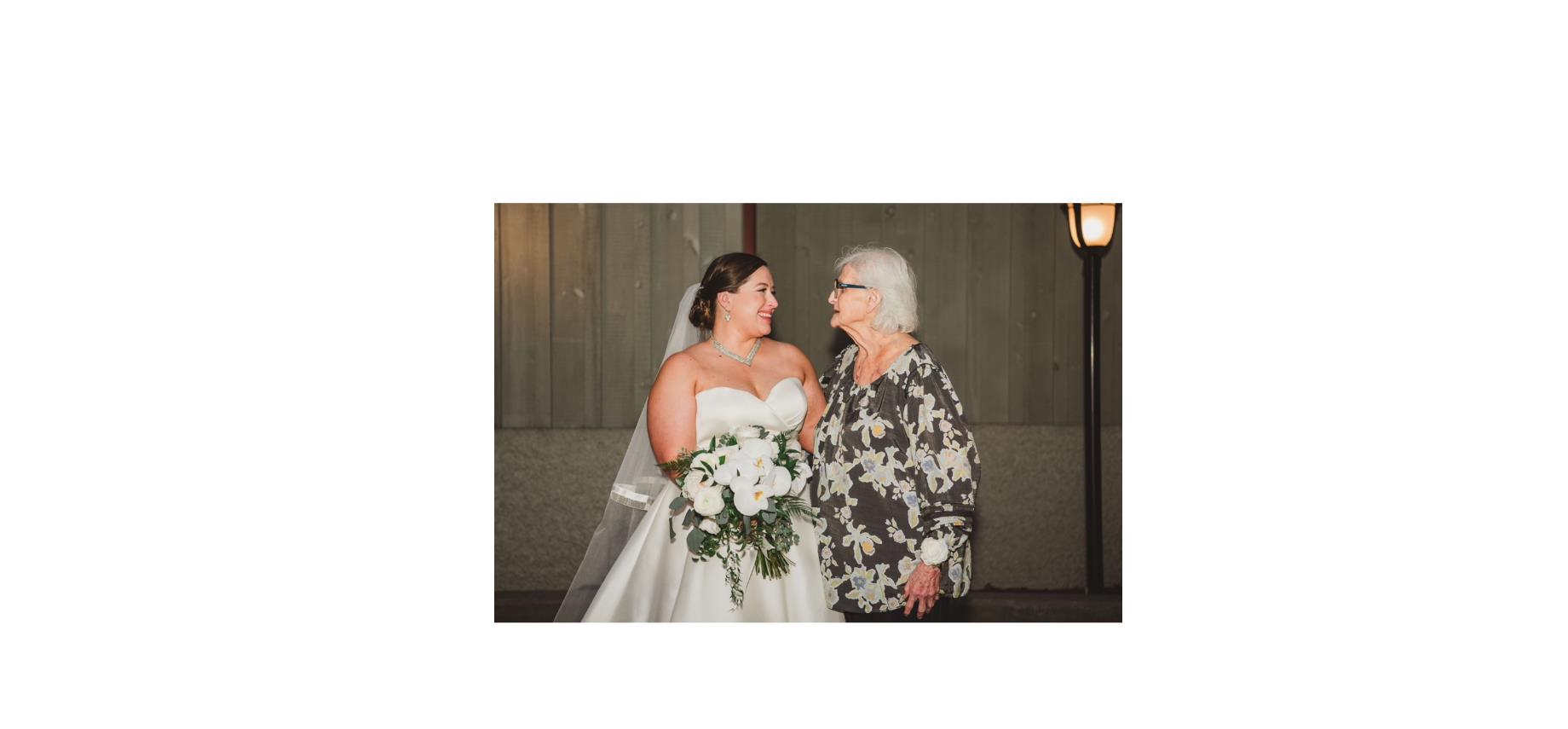 A bride in a white gown smiles at her grandmother, both enjoying a special moment together outside.
