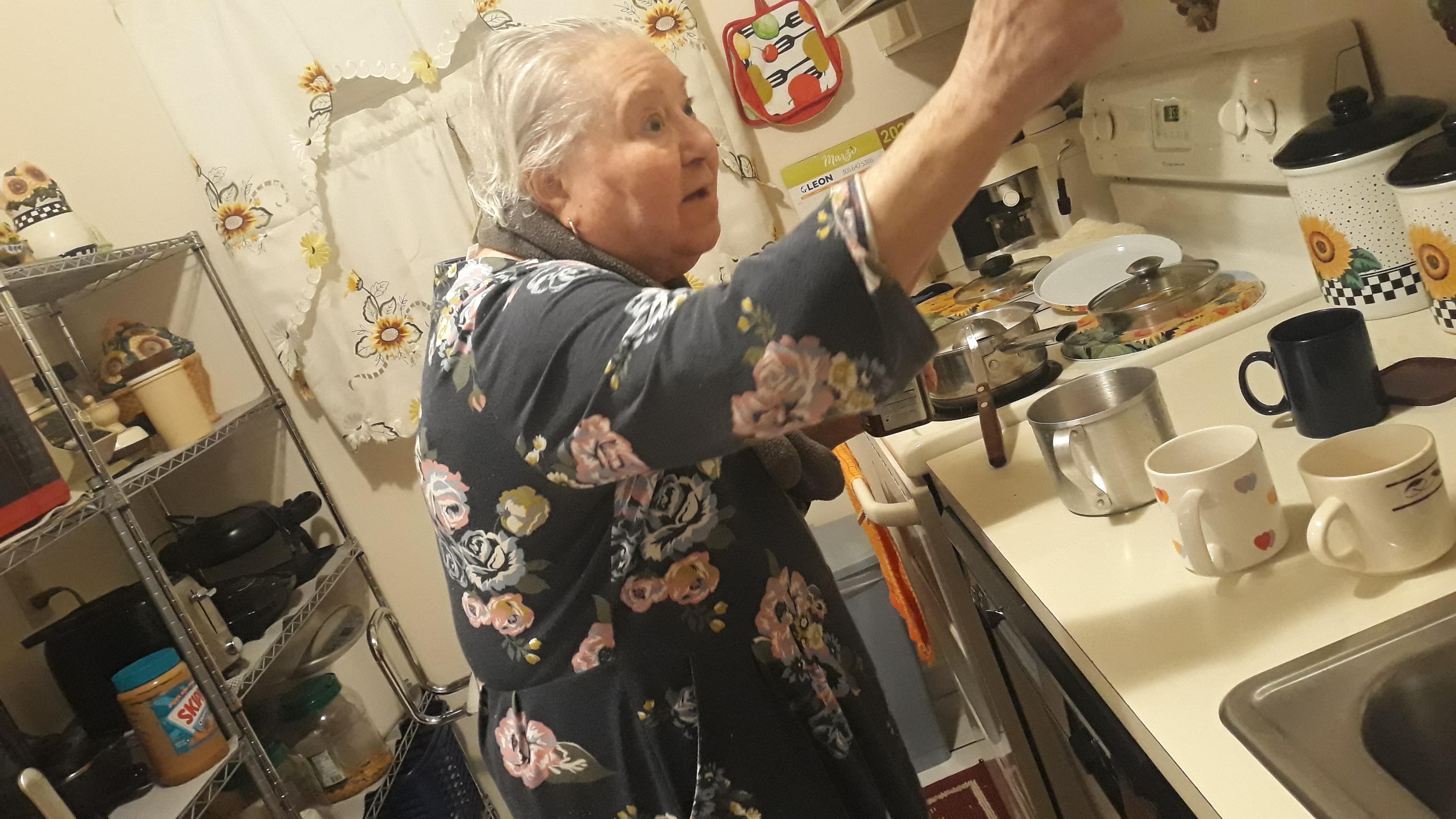 An elderly woman stands in a tidy kitchen, cooking and reminiscing about family traditions.