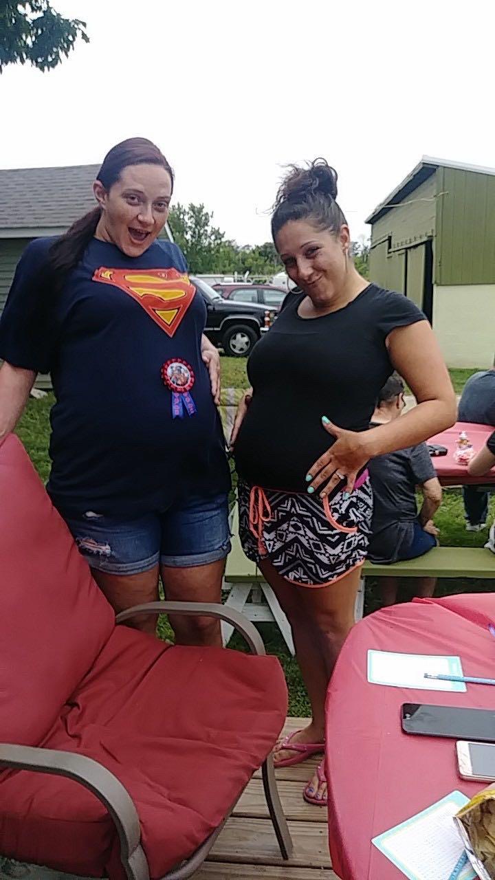 Two friends proudly display their baby bumps at a casual outdoor party surrounded by family.
