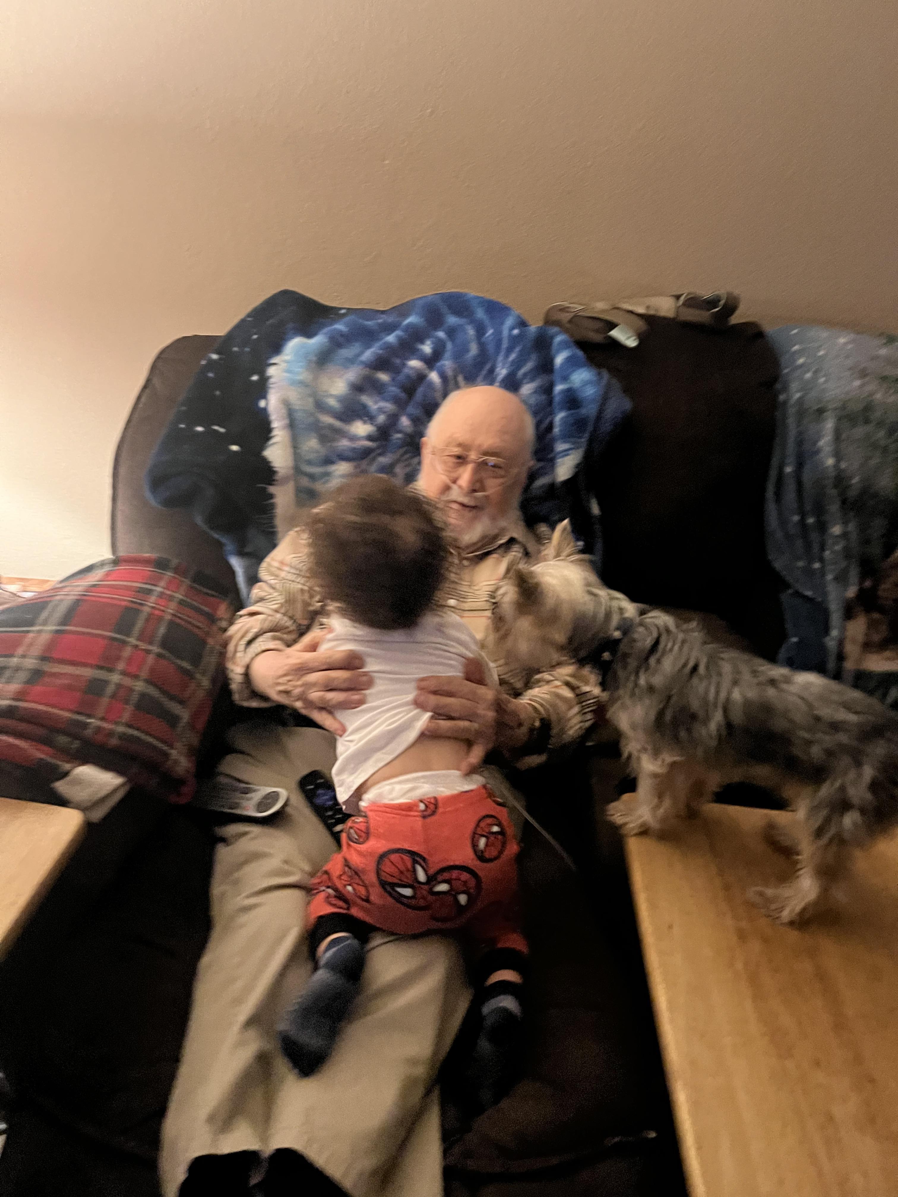 Grandfather sits comfortably on a couch, engaging with a toddler and a small dog.
