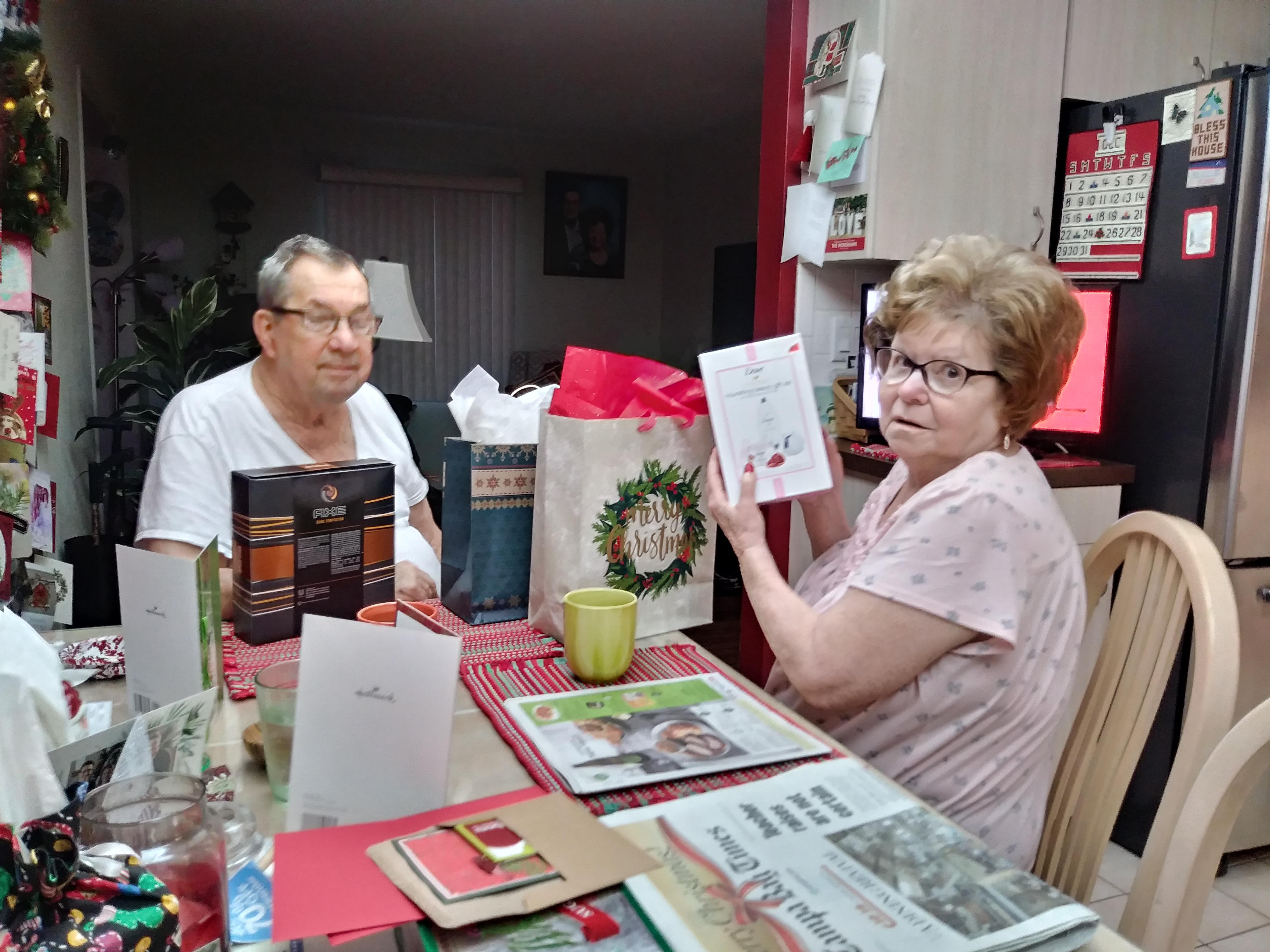 An elderly couple sits together at a dining table, exchanging gifts and holiday greetings.