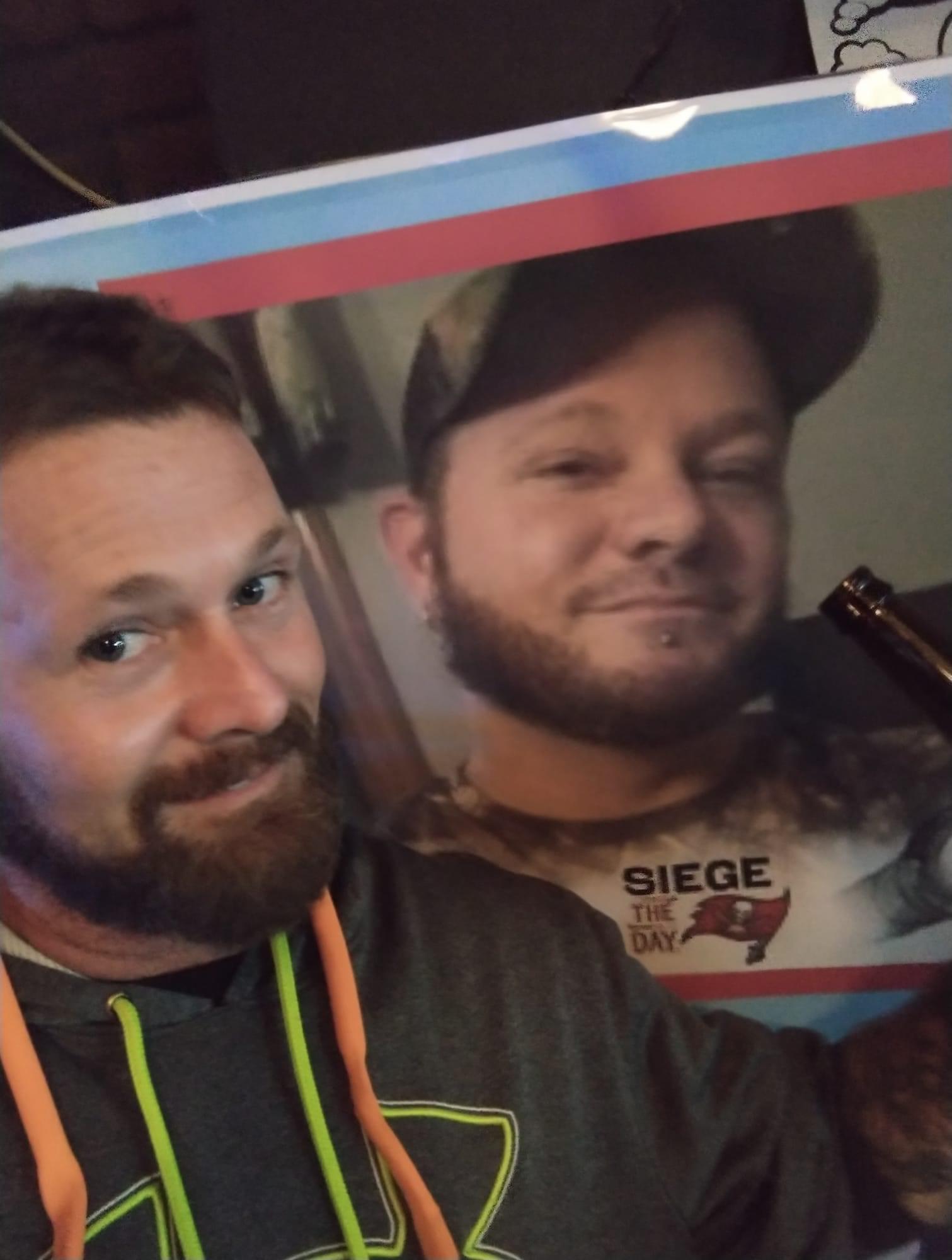 Individual smiles and poses alongside a large portrait at a lively gathering during the evening.