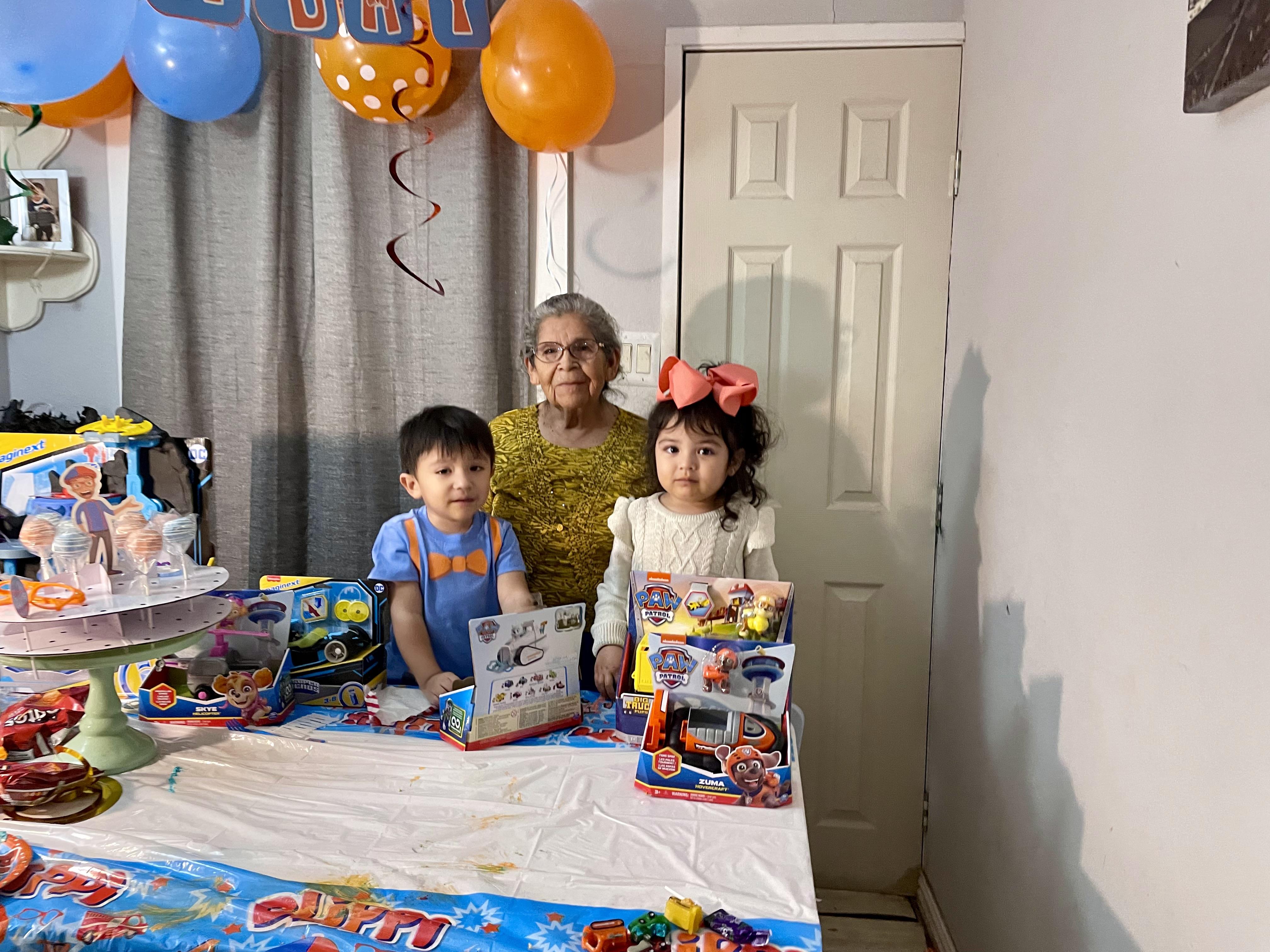 Two children and their grandmother gather around a decorated table filled with toys and treats.