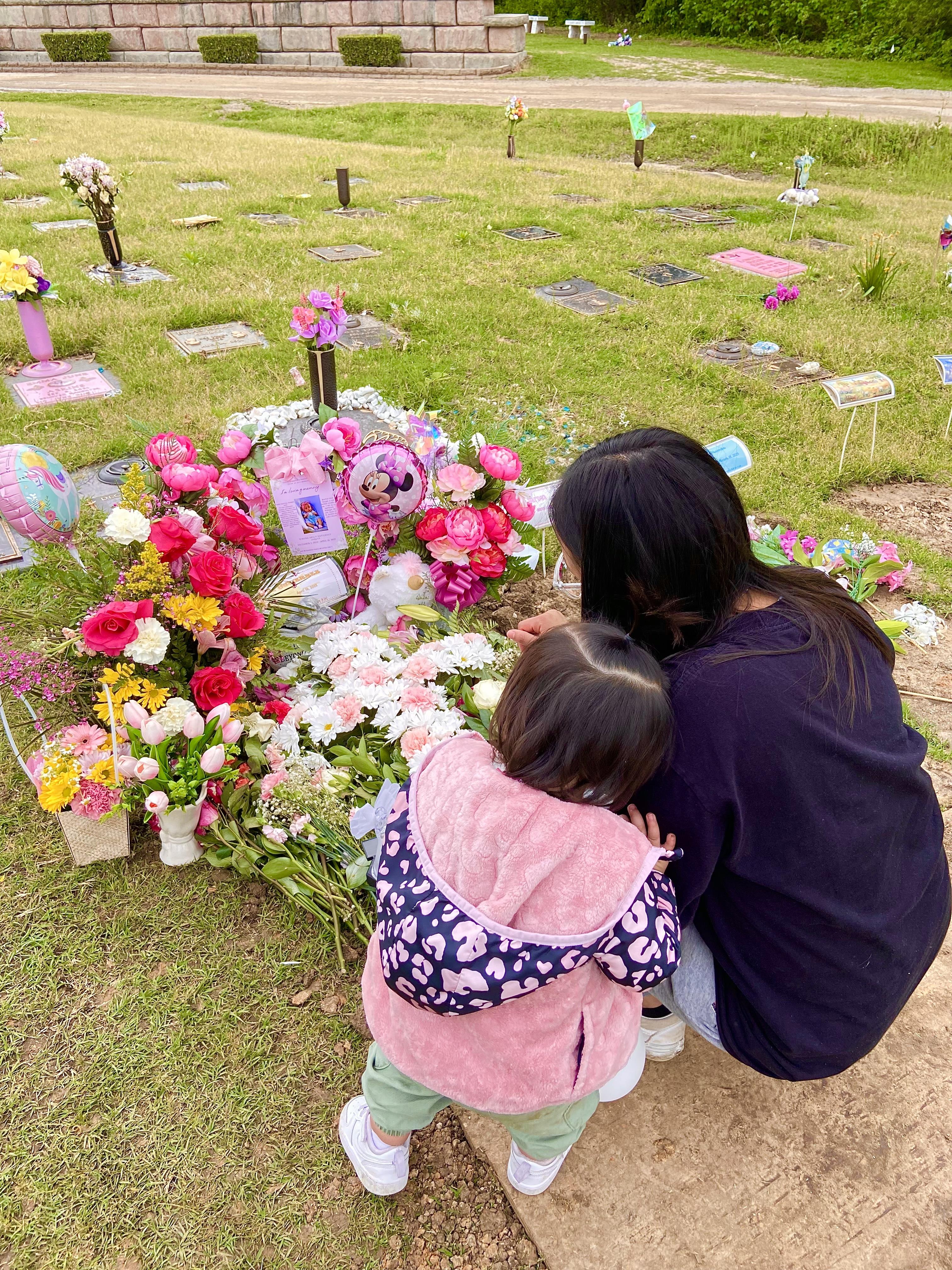 A woman and child pay respects at a grave, surrounded by colorful flowers and remembrance items.