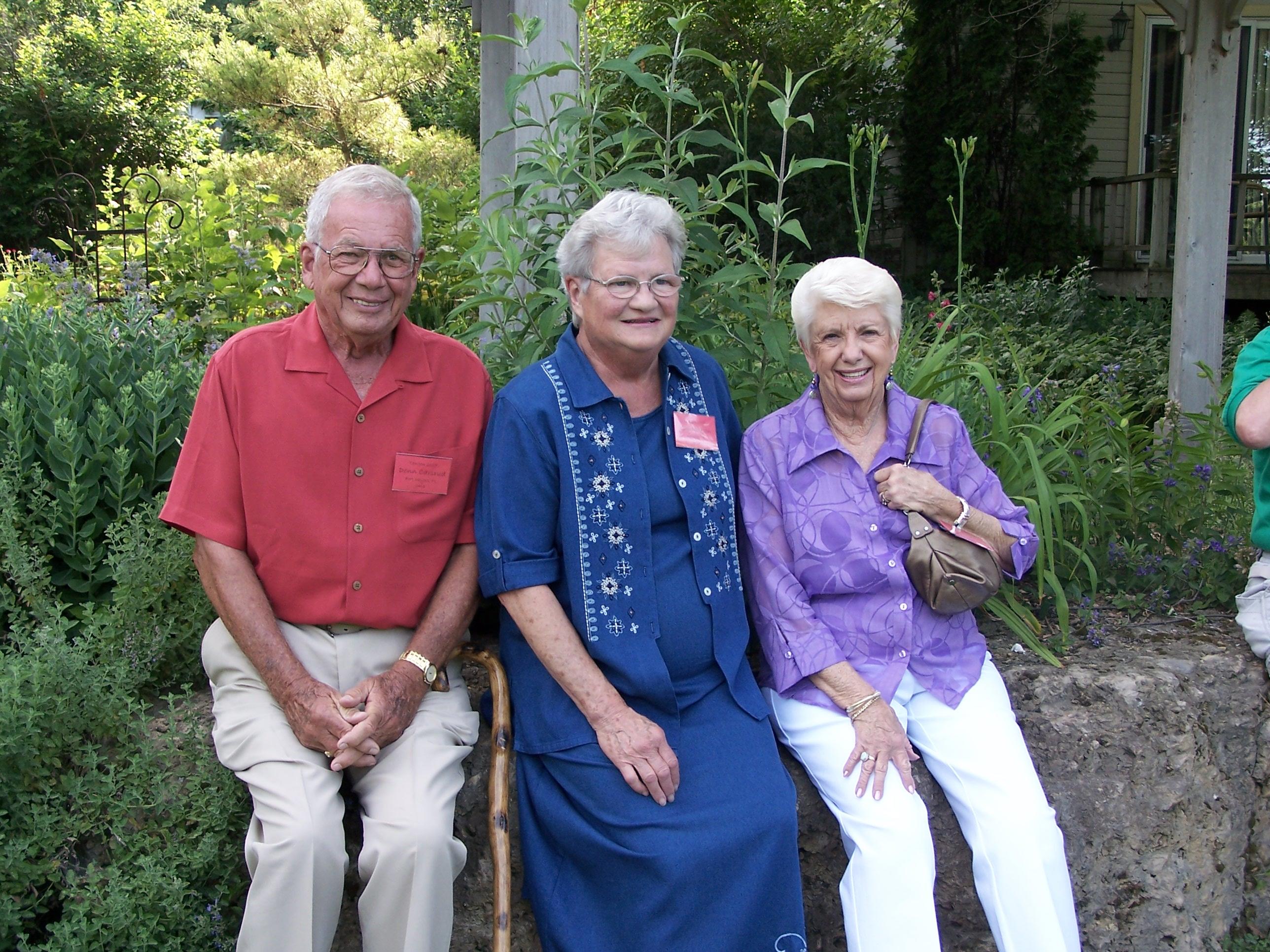 Three elderly individuals sit happily in a lush garden, relishing the warmth of summer.