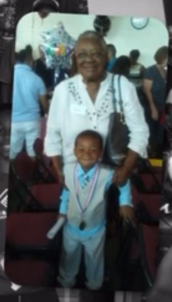 A proud grandmother stands beside her grandson, who is wearing a formal outfit and medal.