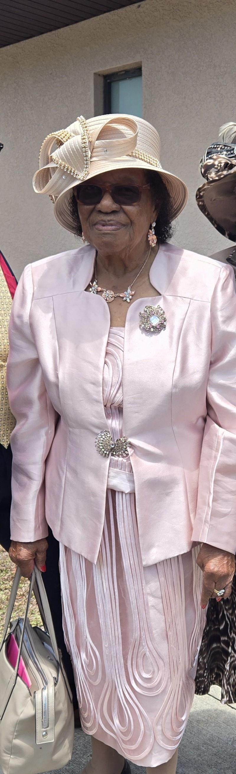 A woman dressed in a sophisticated light pink suit and hat stands proudly at a gathering.