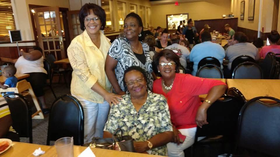 Four women pose happily together at a restaurant, surrounded by family and friends enjoying dinner.