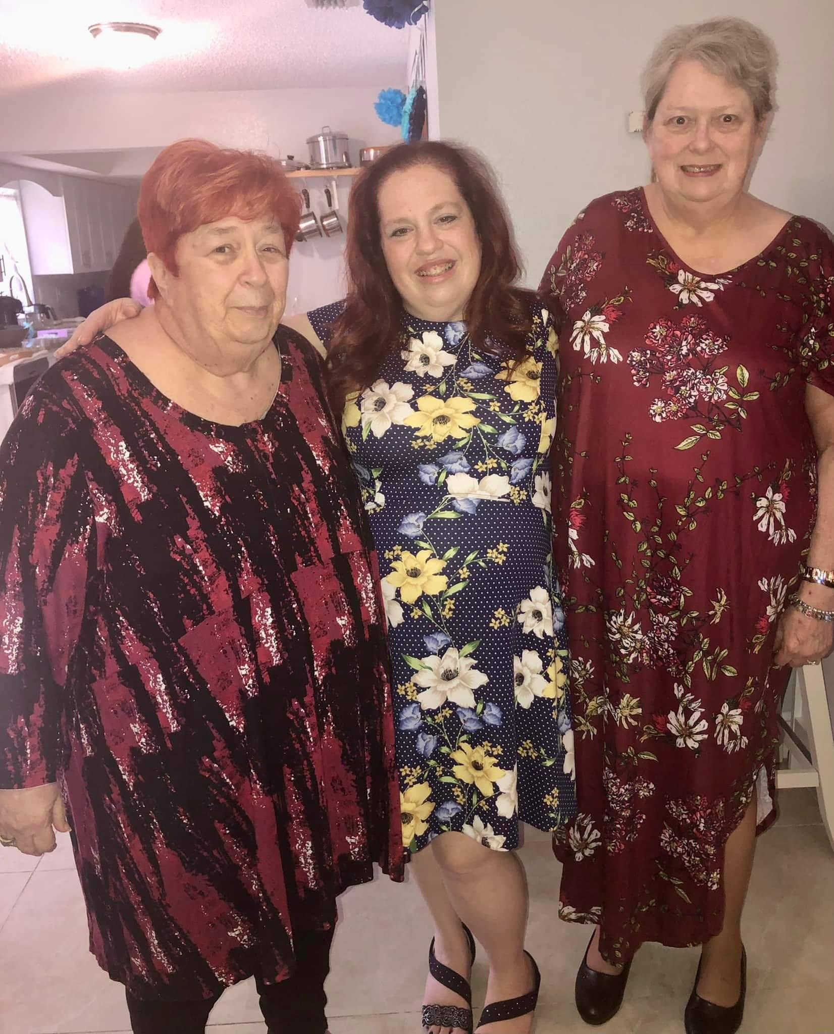 Three women pose happily in colorful dresses during a family gathering in a welcoming home.
