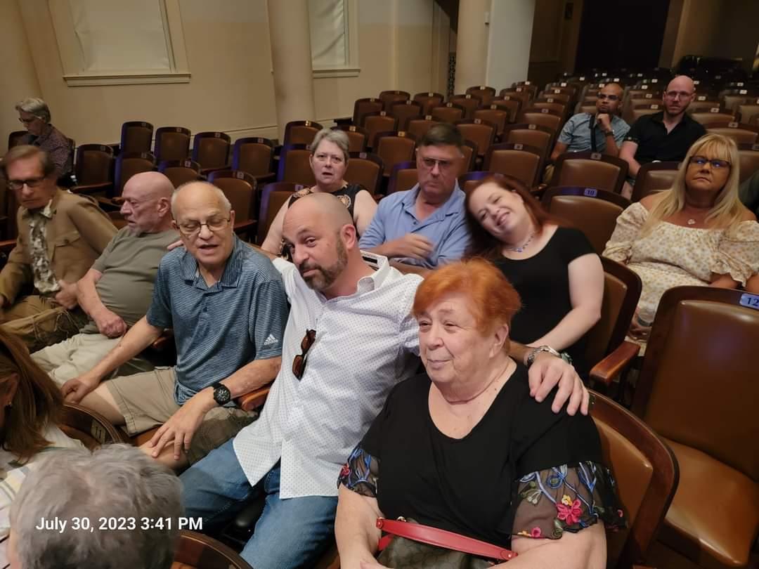 Audience members are seated in a theater, engaged in conversation before a performance.