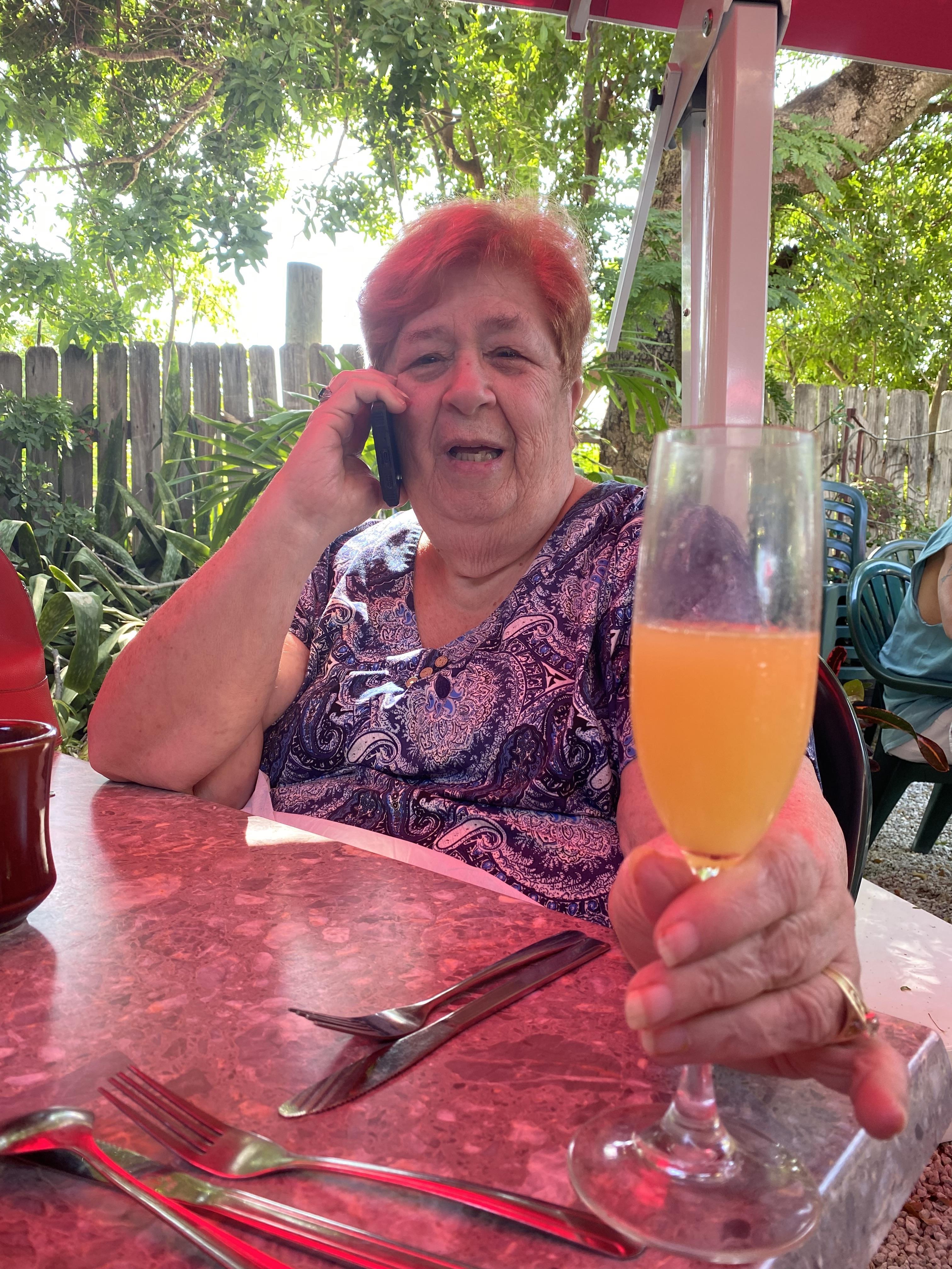 Woman smiles as she chats on the phone, holding a drink at a vibrant outdoor restaurant.
