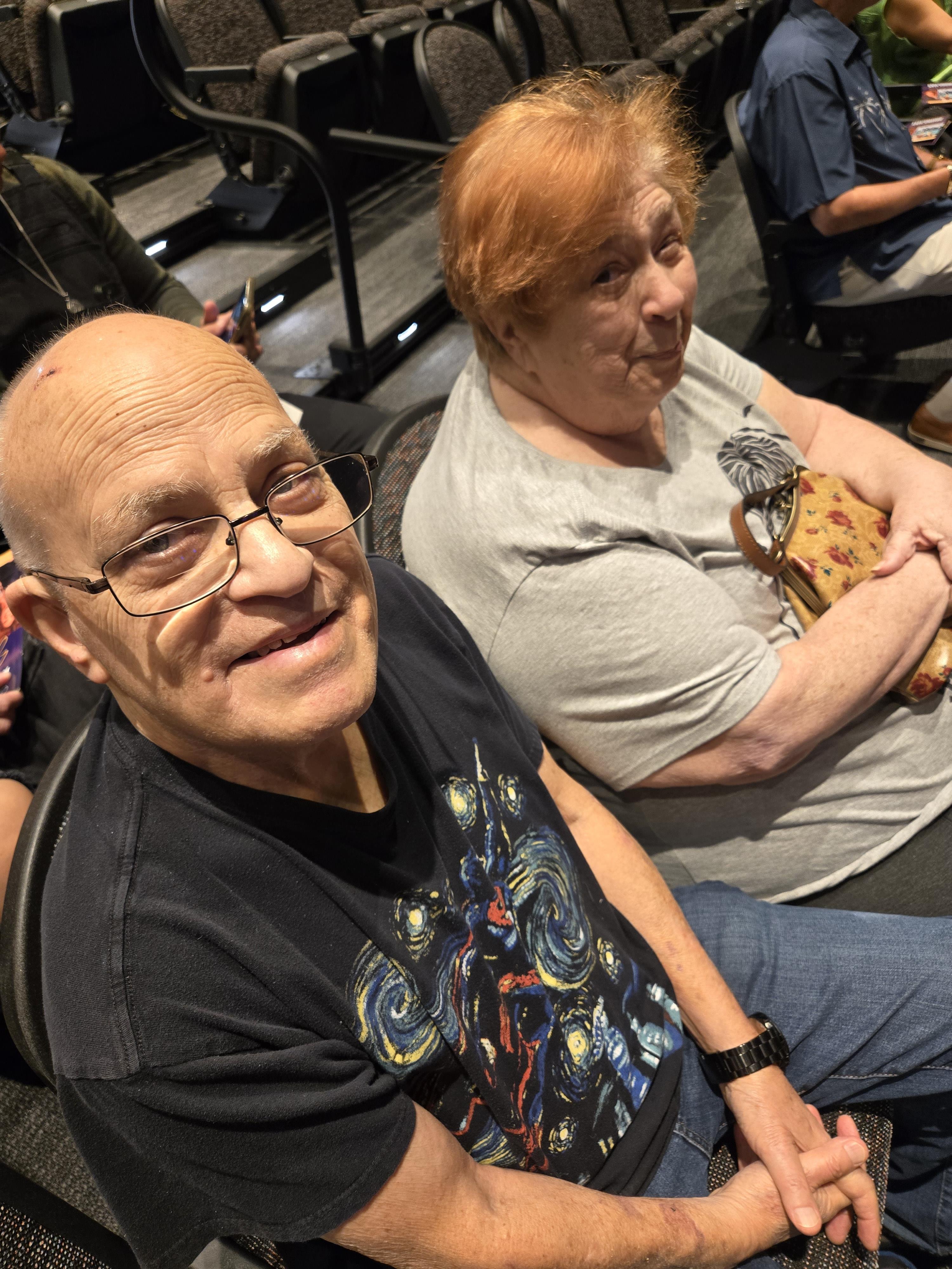 An elderly couple sit closely together in a theater, both smiling and enjoying a community event.