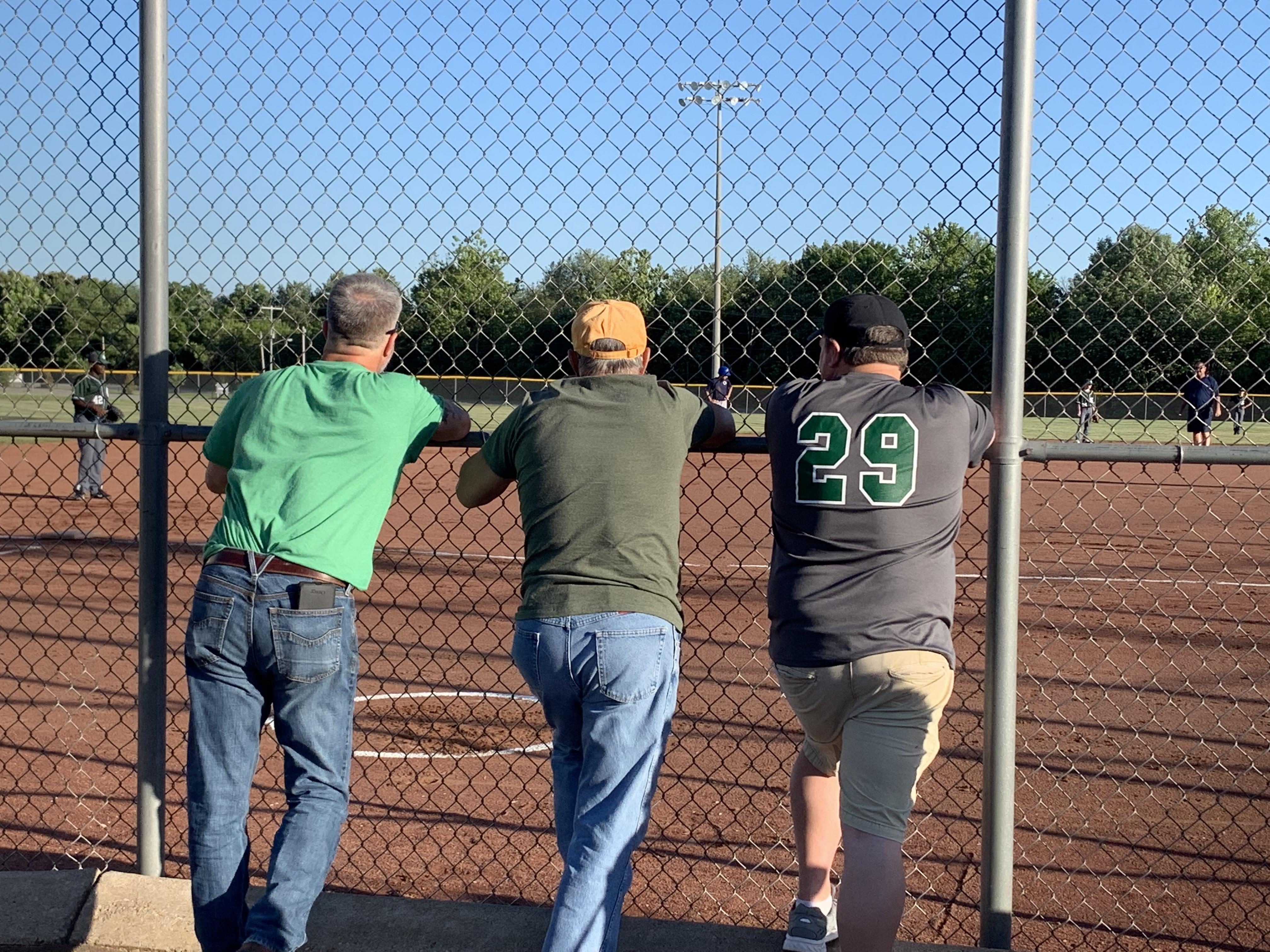 Three men lean against a fence, watching a youth baseball game in a park.