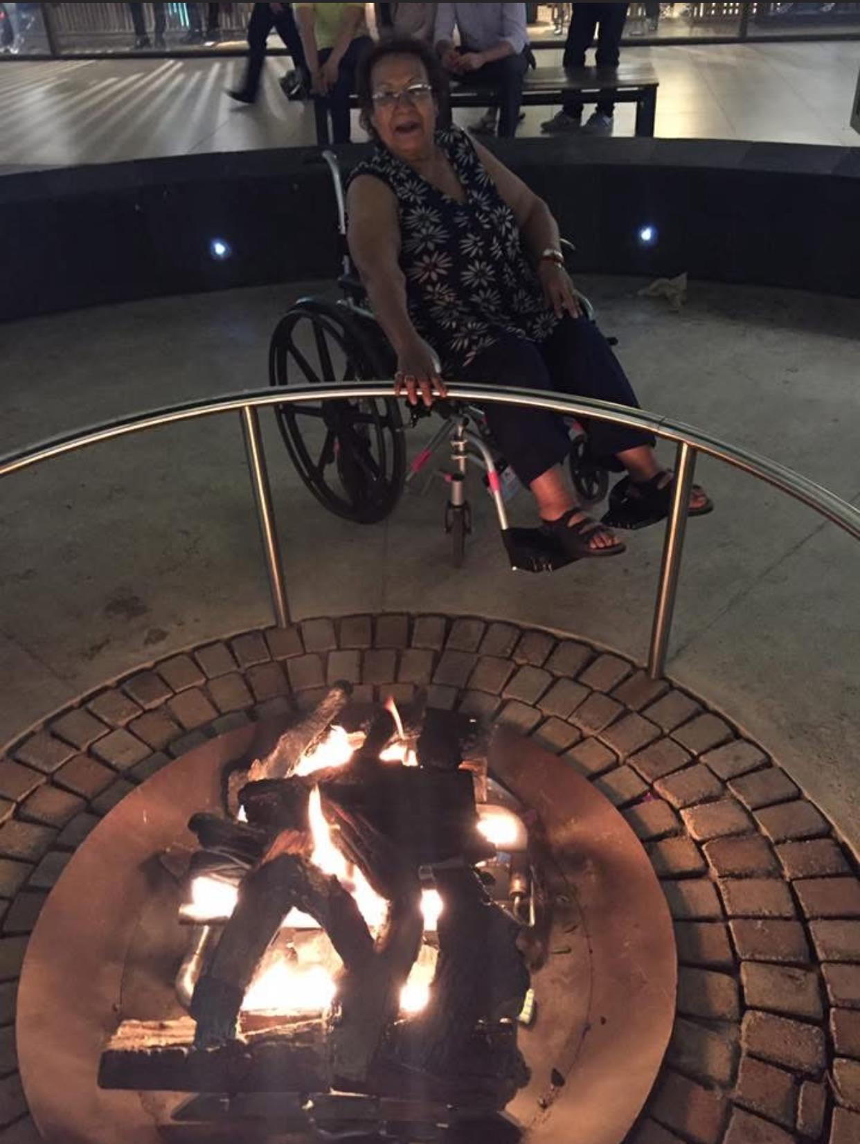 A person in a wheelchair sits by a campfire, surrounded by a lively outdoor gathering atmosphere.