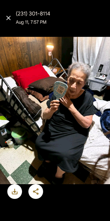 An elderly woman sits on a bed, holding a mirror and surrounded by everyday items in a room.