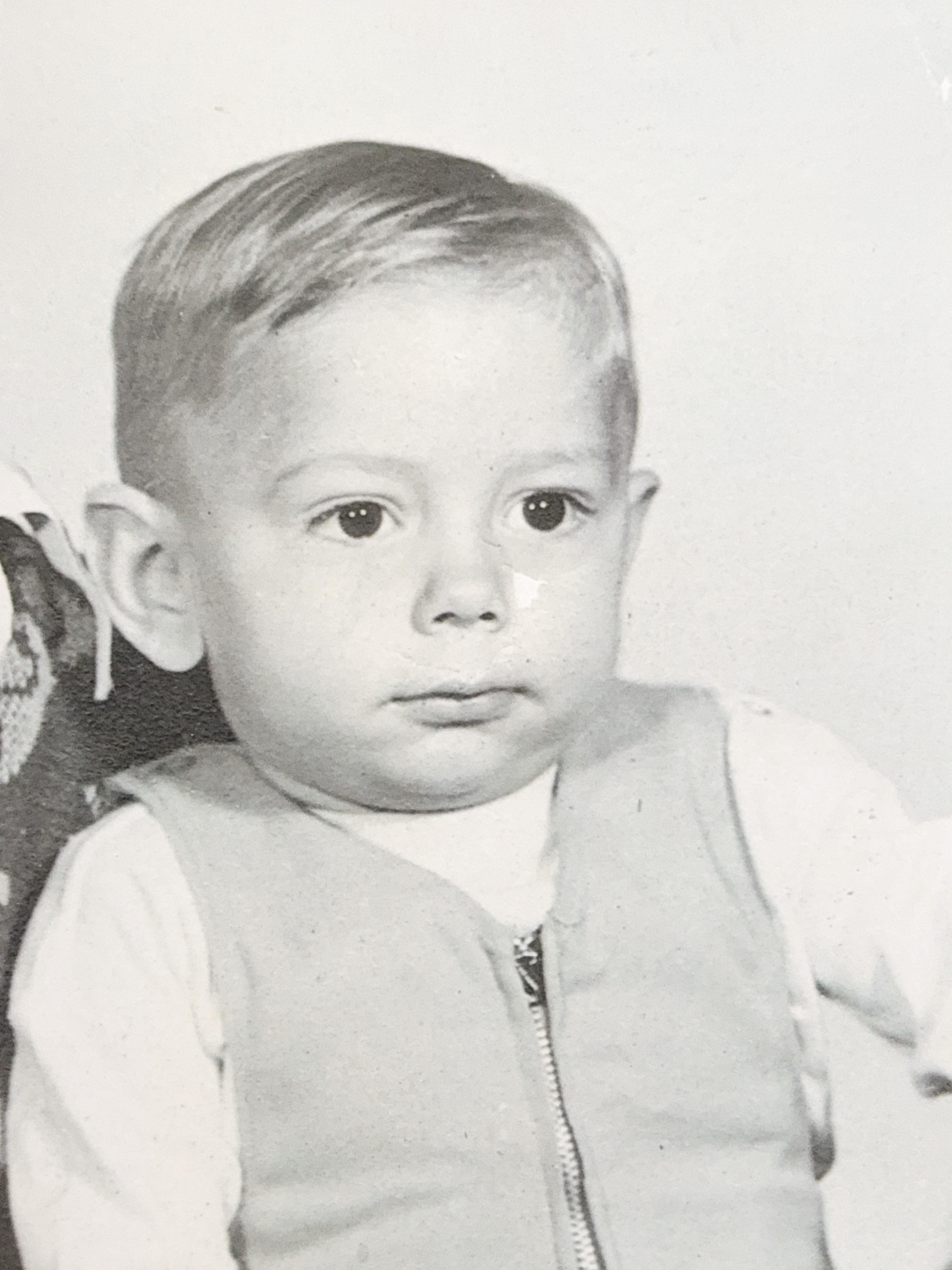 A young child with light hair sits in a vintage studio, wearing a vest, looking curious.
