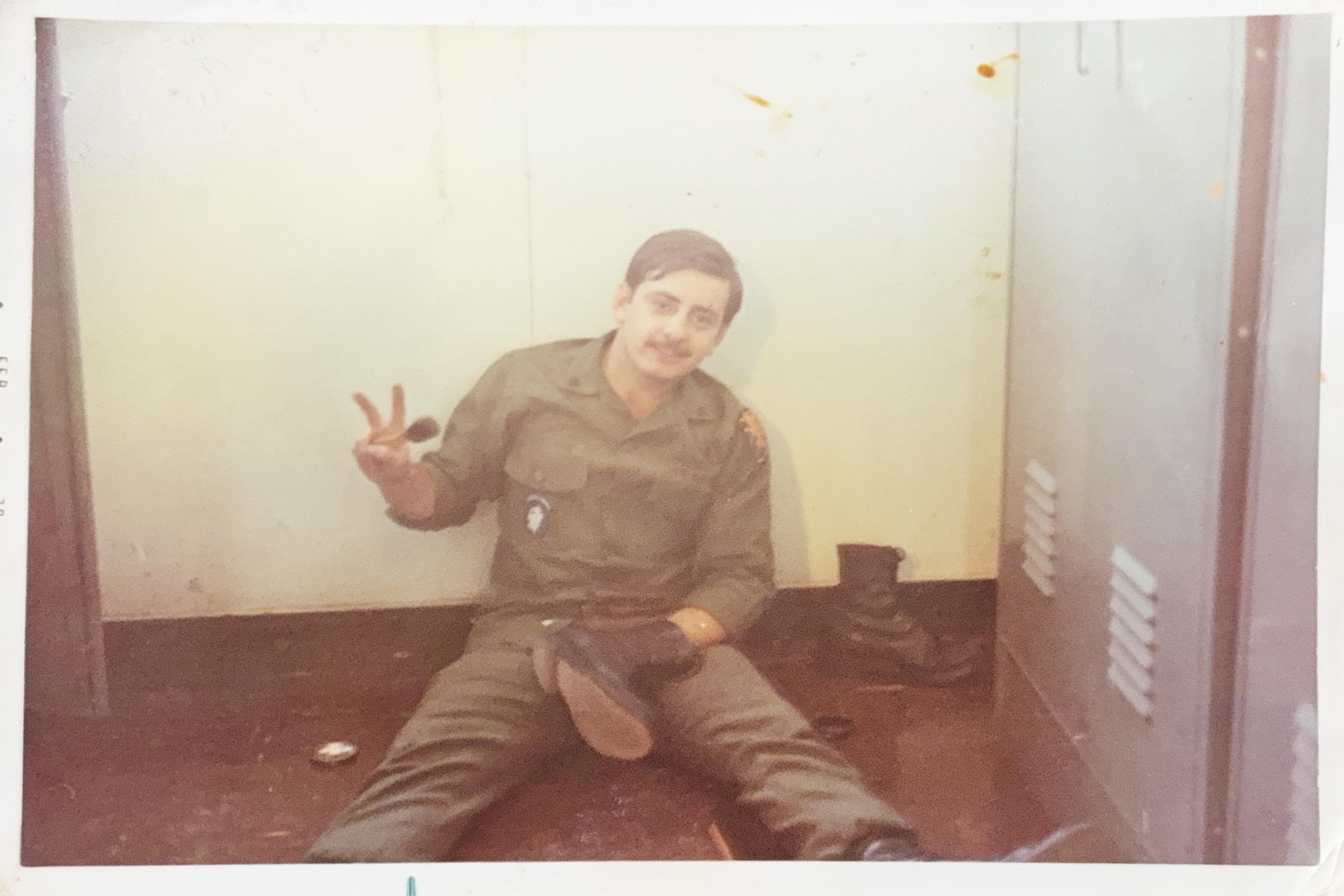 A smiling soldier dressed in uniform sits casually on the floor, making a peace sign with his hand.