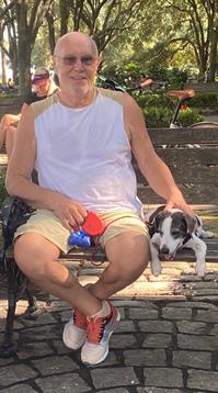 A man sits on a bench with a small dog beside him, enjoying a sunny day in the park.