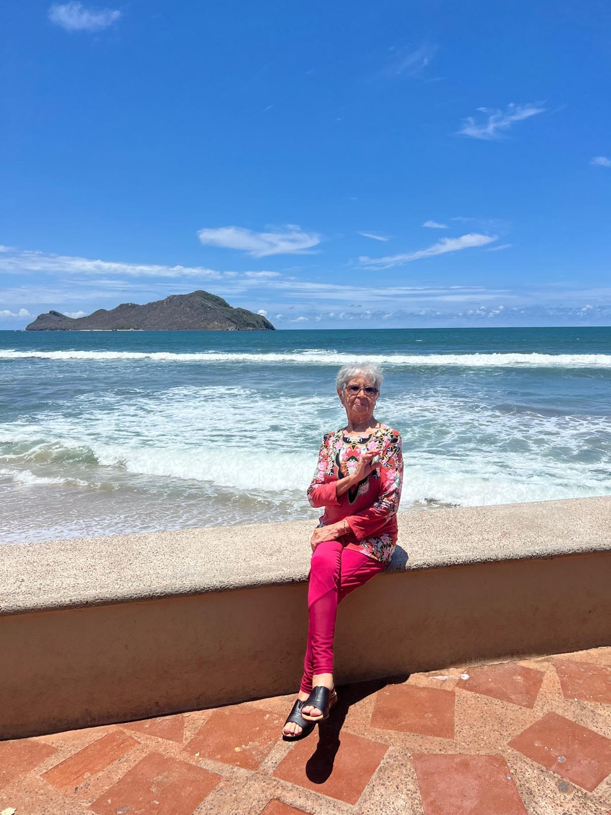 A woman sits relaxed on a seawall, soaking up the sun by the ocean with waves rolling in.