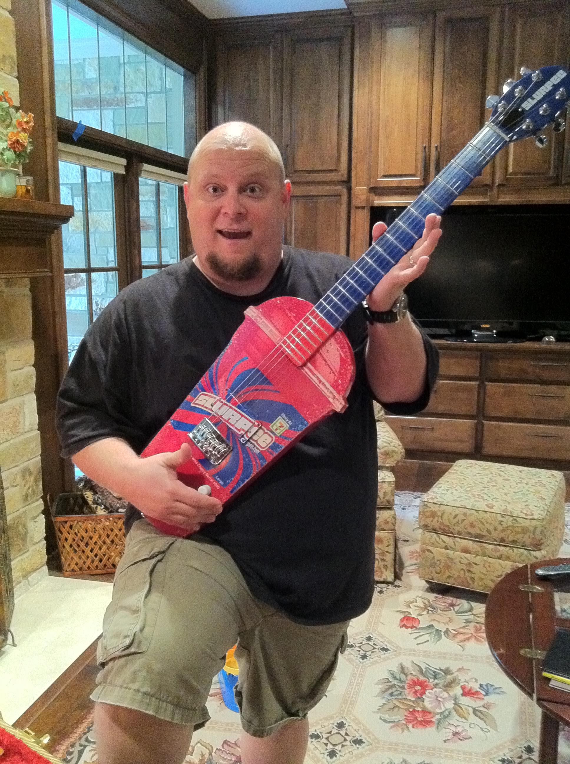 A man poses excitedly with a vibrant handmade guitar in a cozy room filled with wooden furniture.