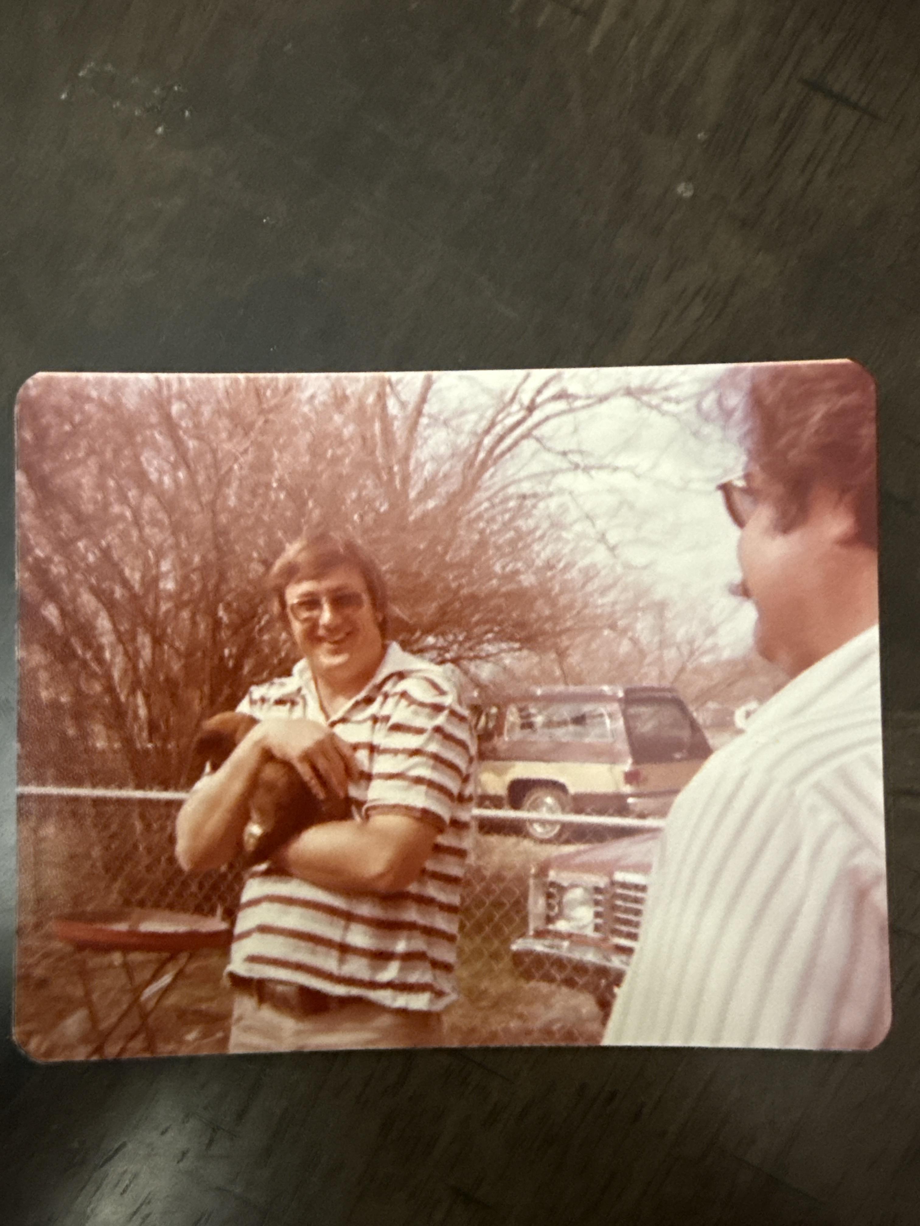 Two men engage in a lively conversation outside. One man holds a small dog close while smiling.