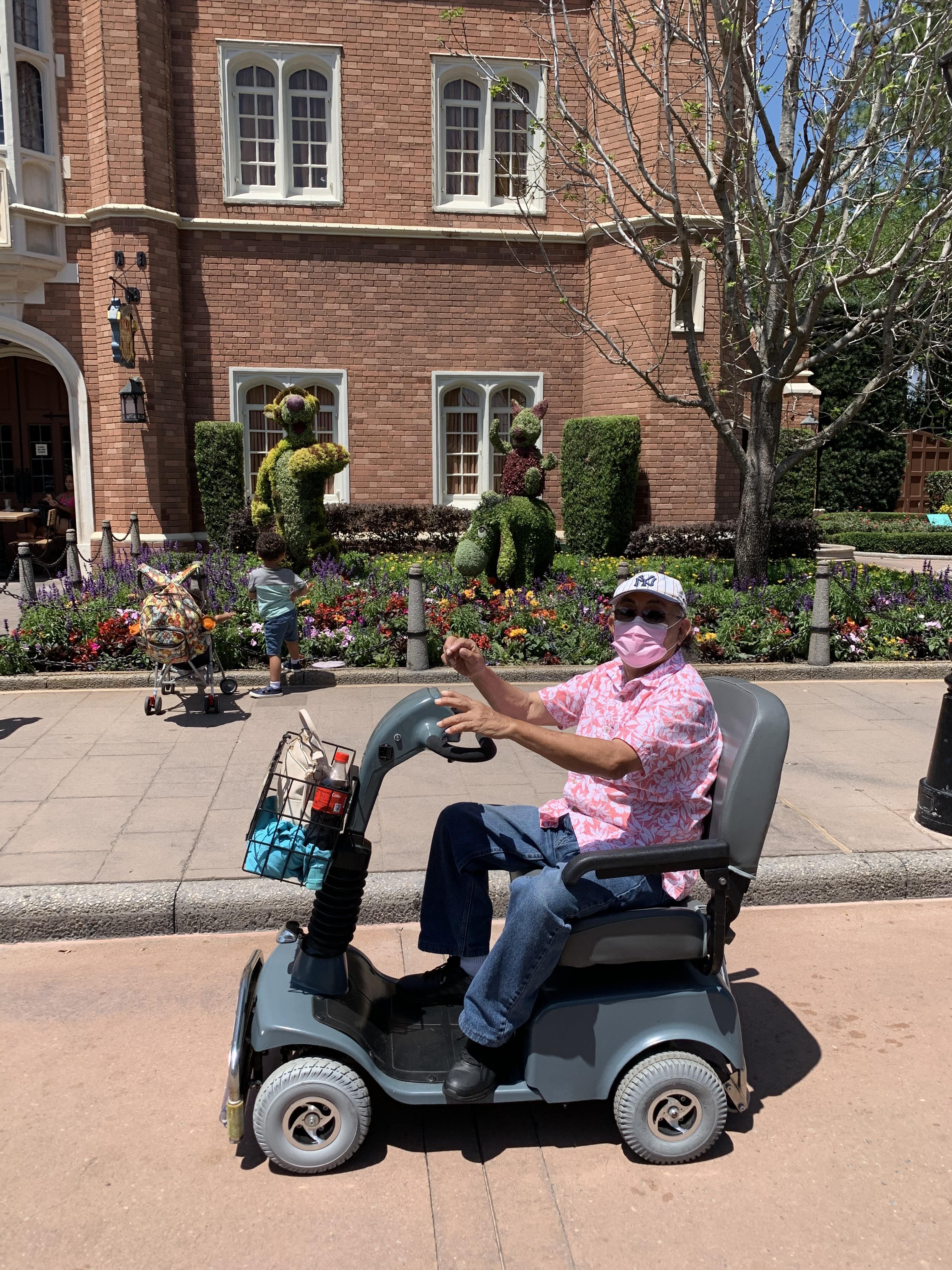 Senior man on scooter smiles at camera while vibrant flowers and lively families surround him.