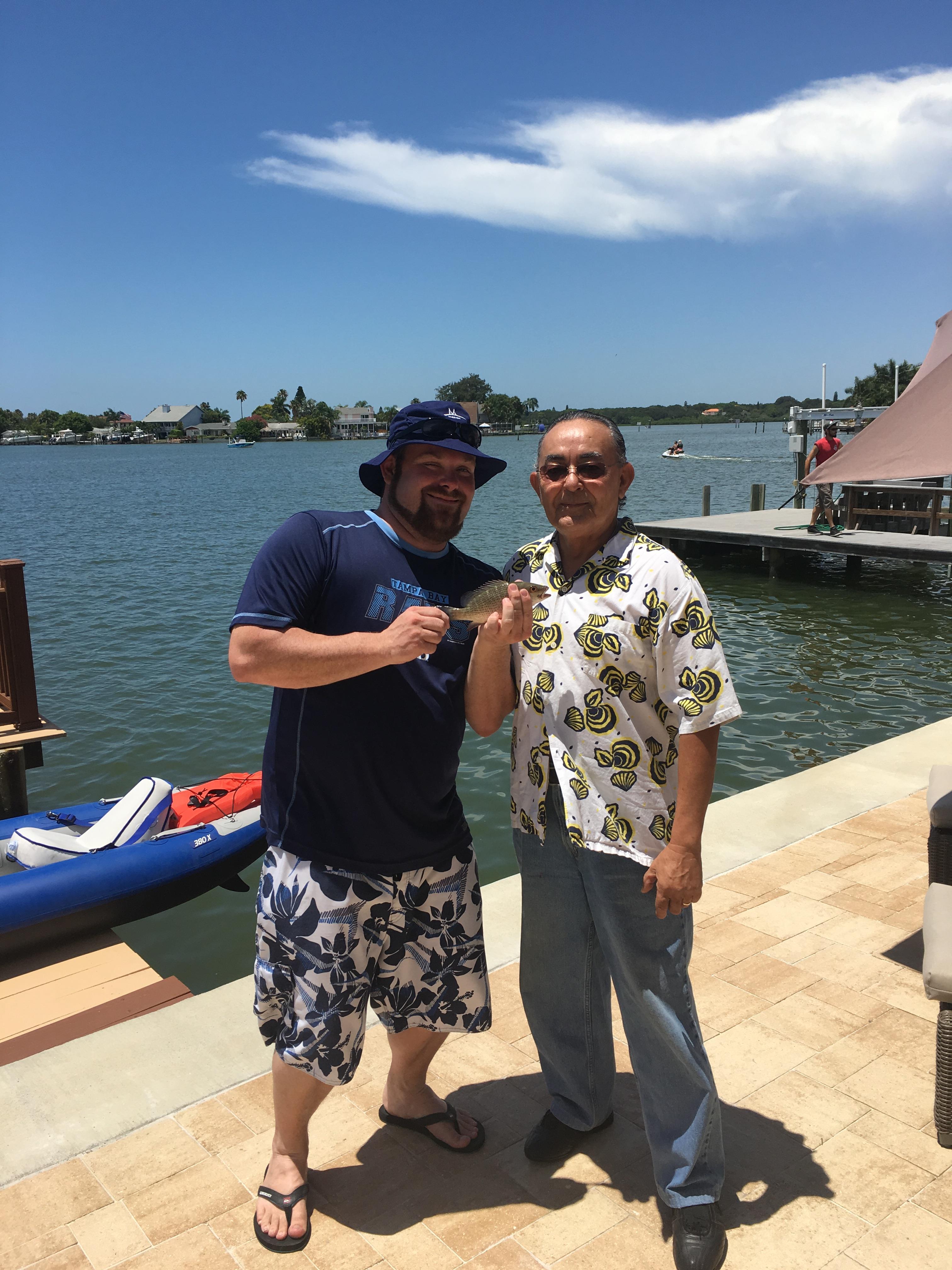 Two men stand by the water, holding a fish, enjoying a sunny day on a dock by the marina.