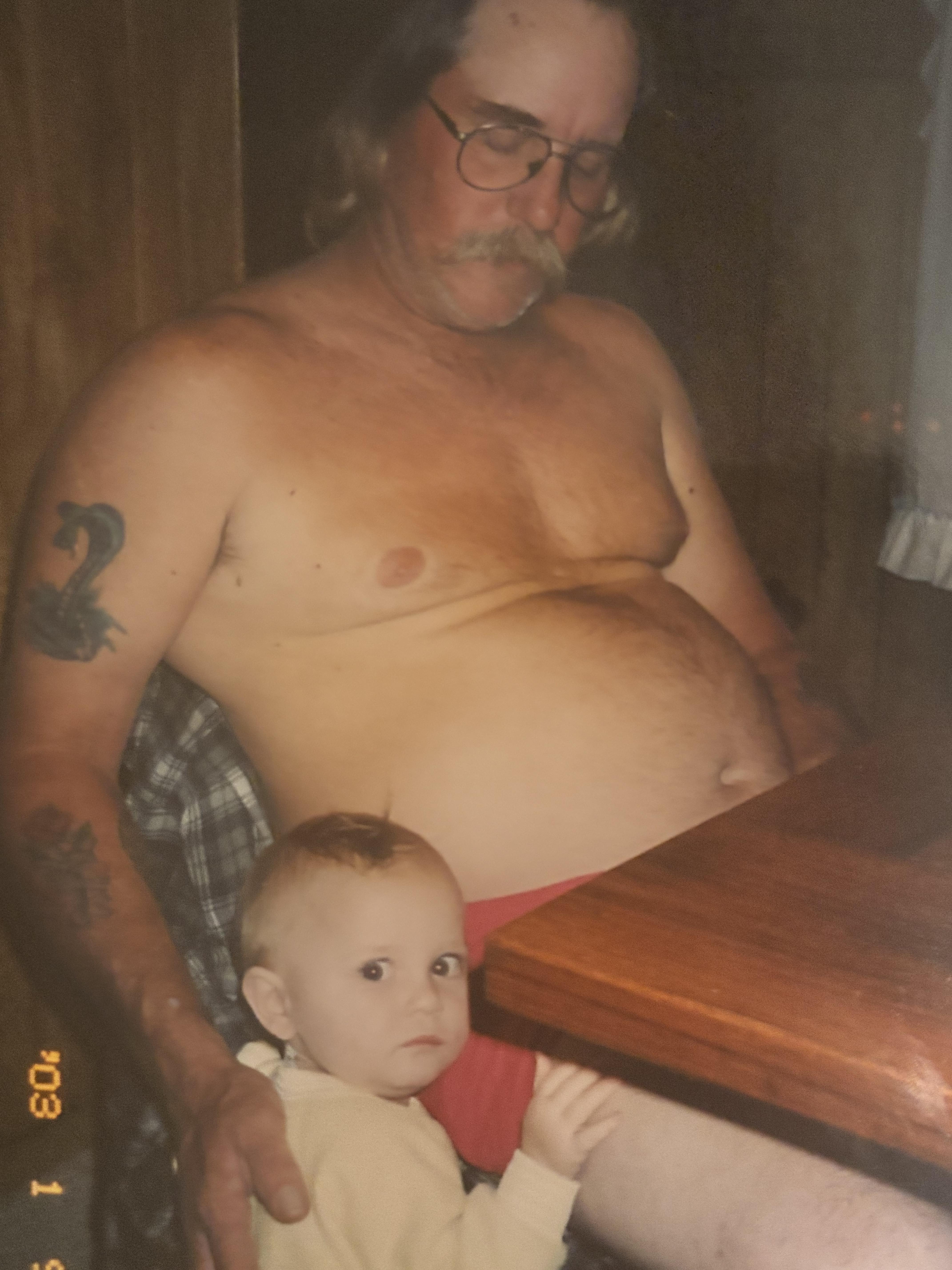 A grandfather sits shirtless at a table while his grandchild curiously watches him.