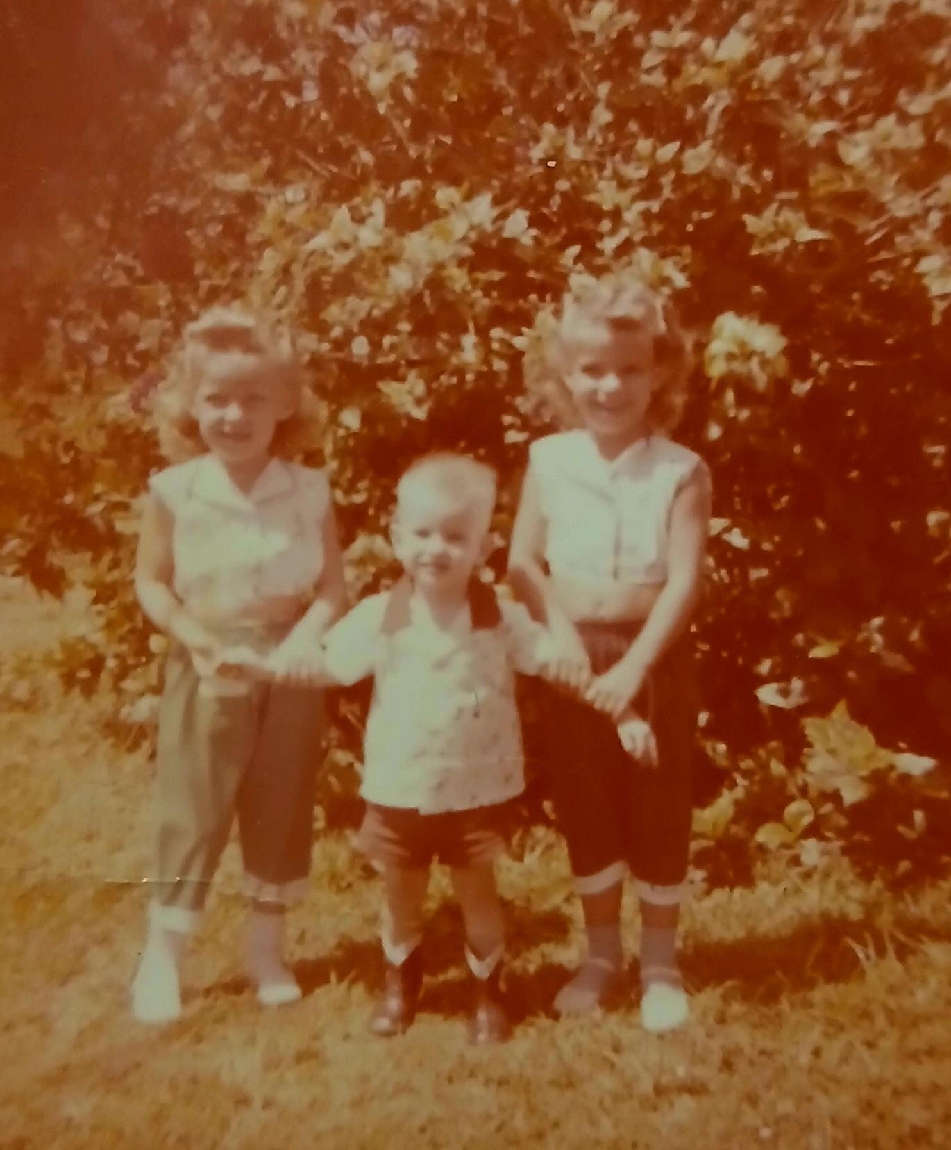 Three children stand together in a garden, smiling and enjoying a sunny day.
