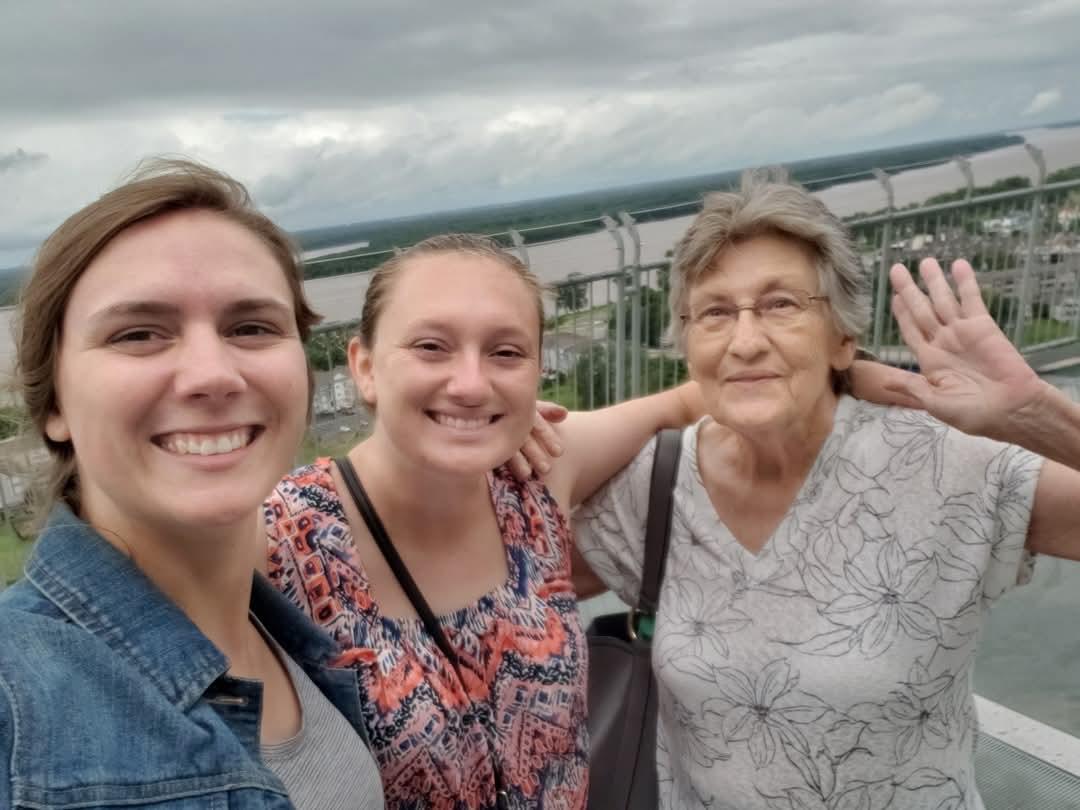 Three women smile and pose for a selfie while admiring the view from a lookout on a cloudy day.