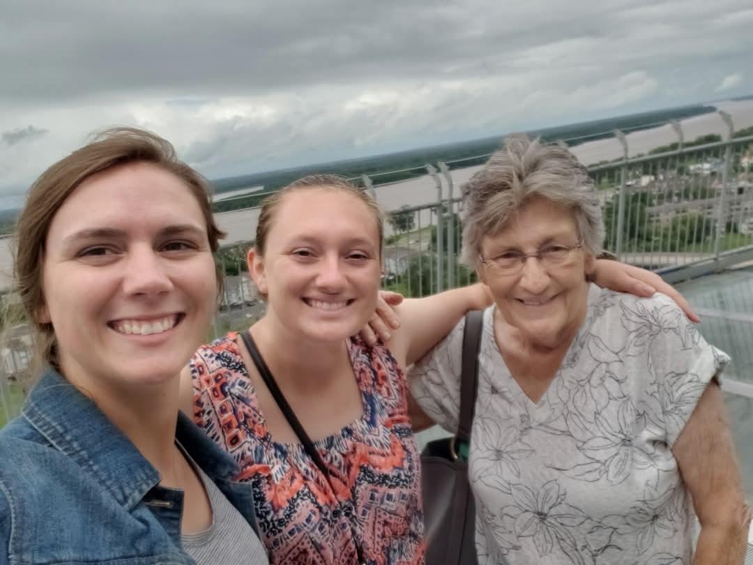 Three women smile together at a high vantage point, enjoying their time on a cloudy afternoon.