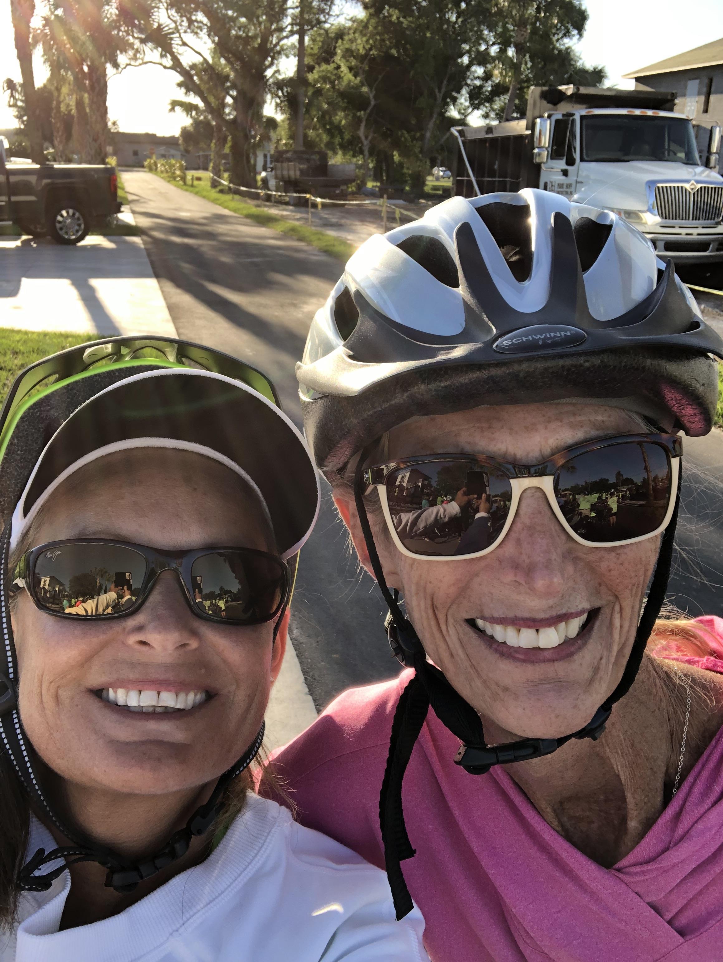 Two women smile brightly while riding bicycles on a scenic coastal road surrounded by palm trees.