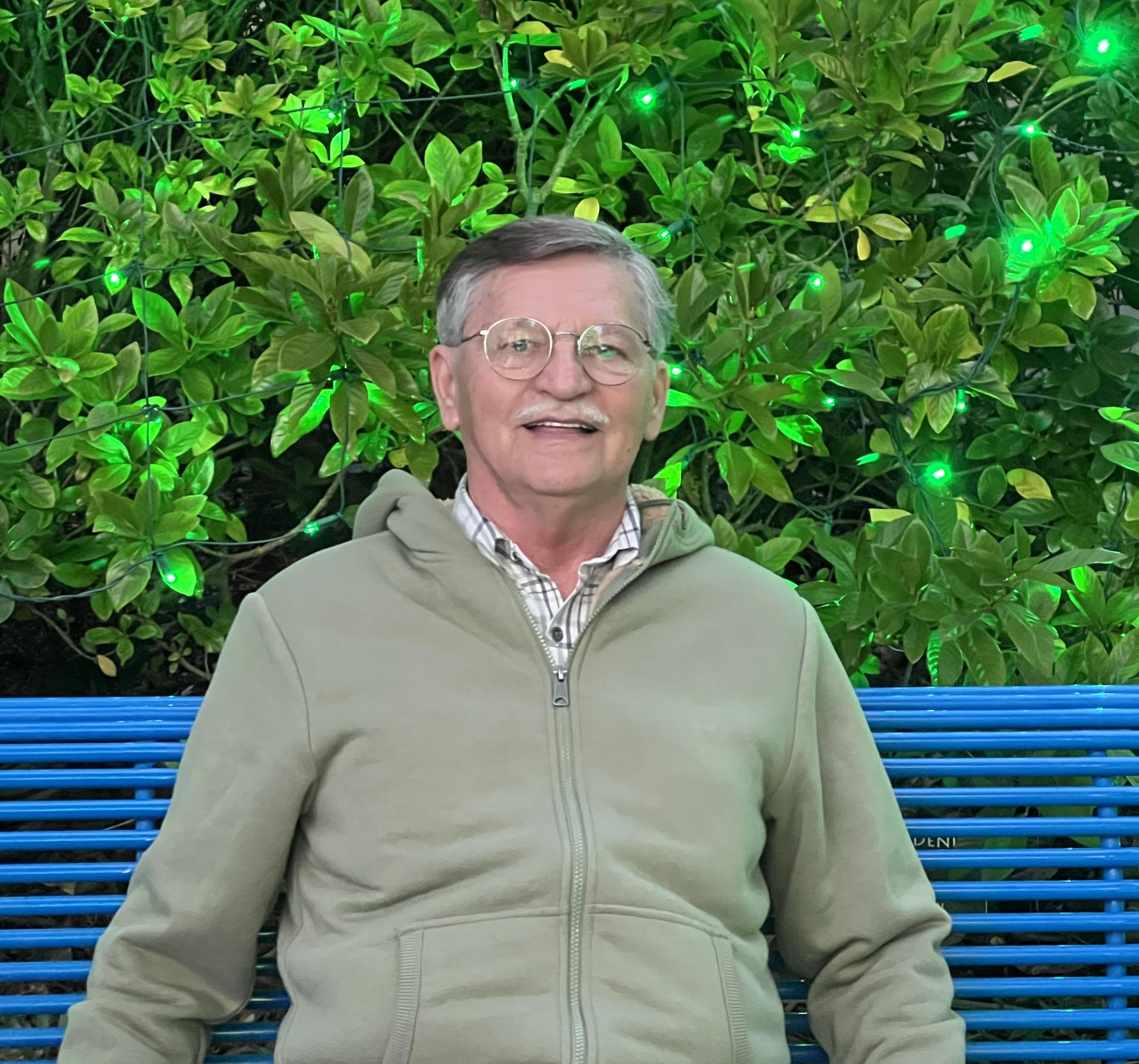 A cheerful senior man sits on a blue bench, surrounded by green plants at twilight.