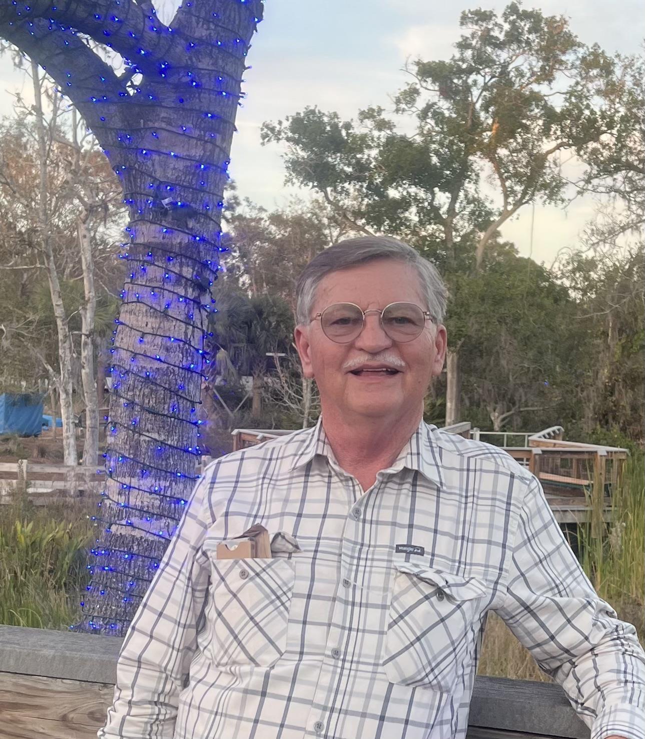 Elderly man stands in front of a lit tree, enjoying a peaceful moment by the water during sunset.