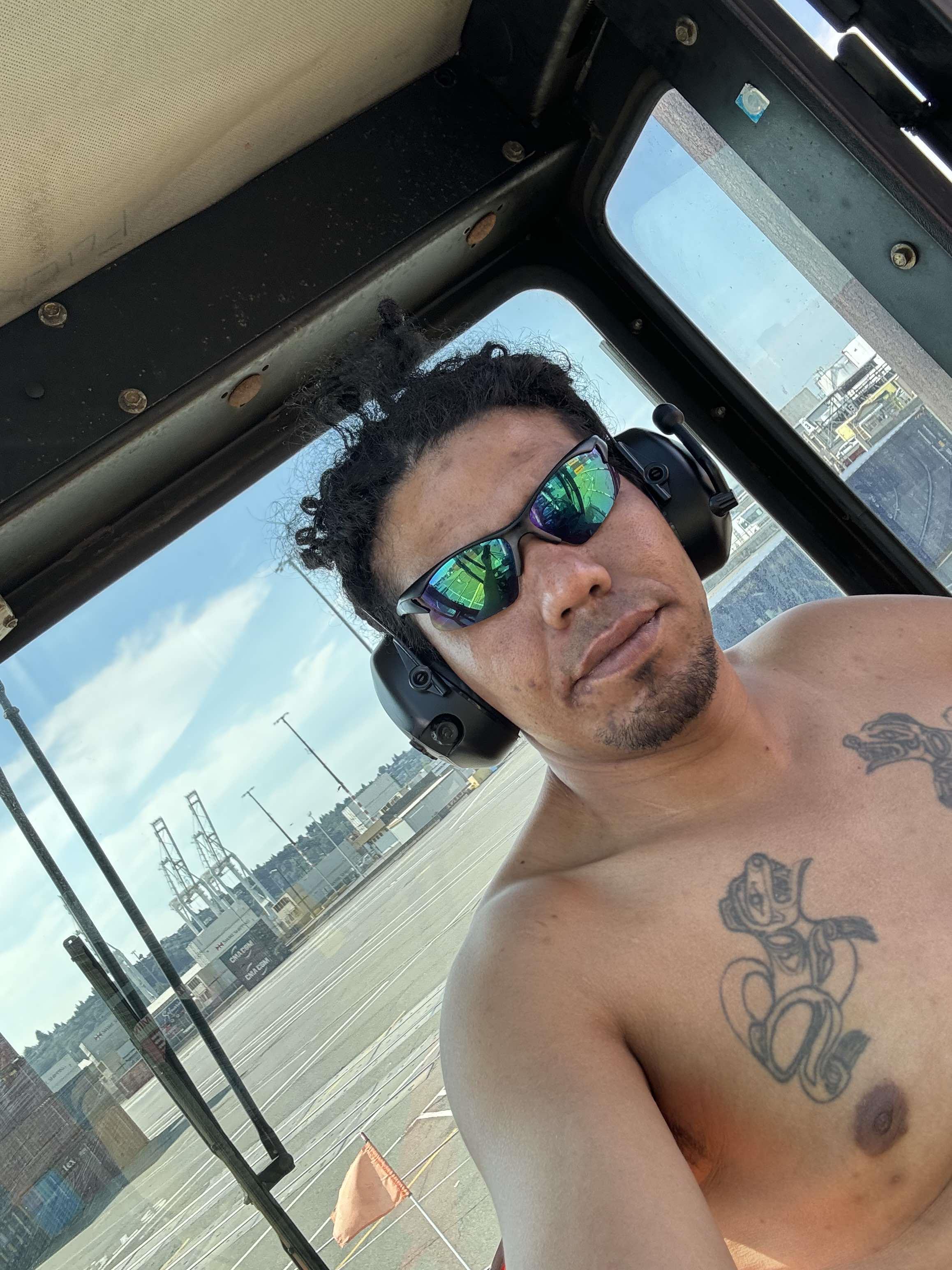 A man relaxes in a construction vehicle at a dock, showing off tattoos and sunglasses during break.