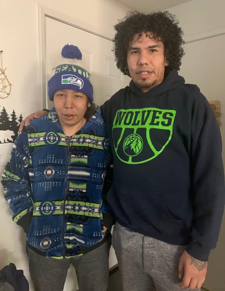 Two friends stand together indoors, casually dressed and smiling warmly at the camera.