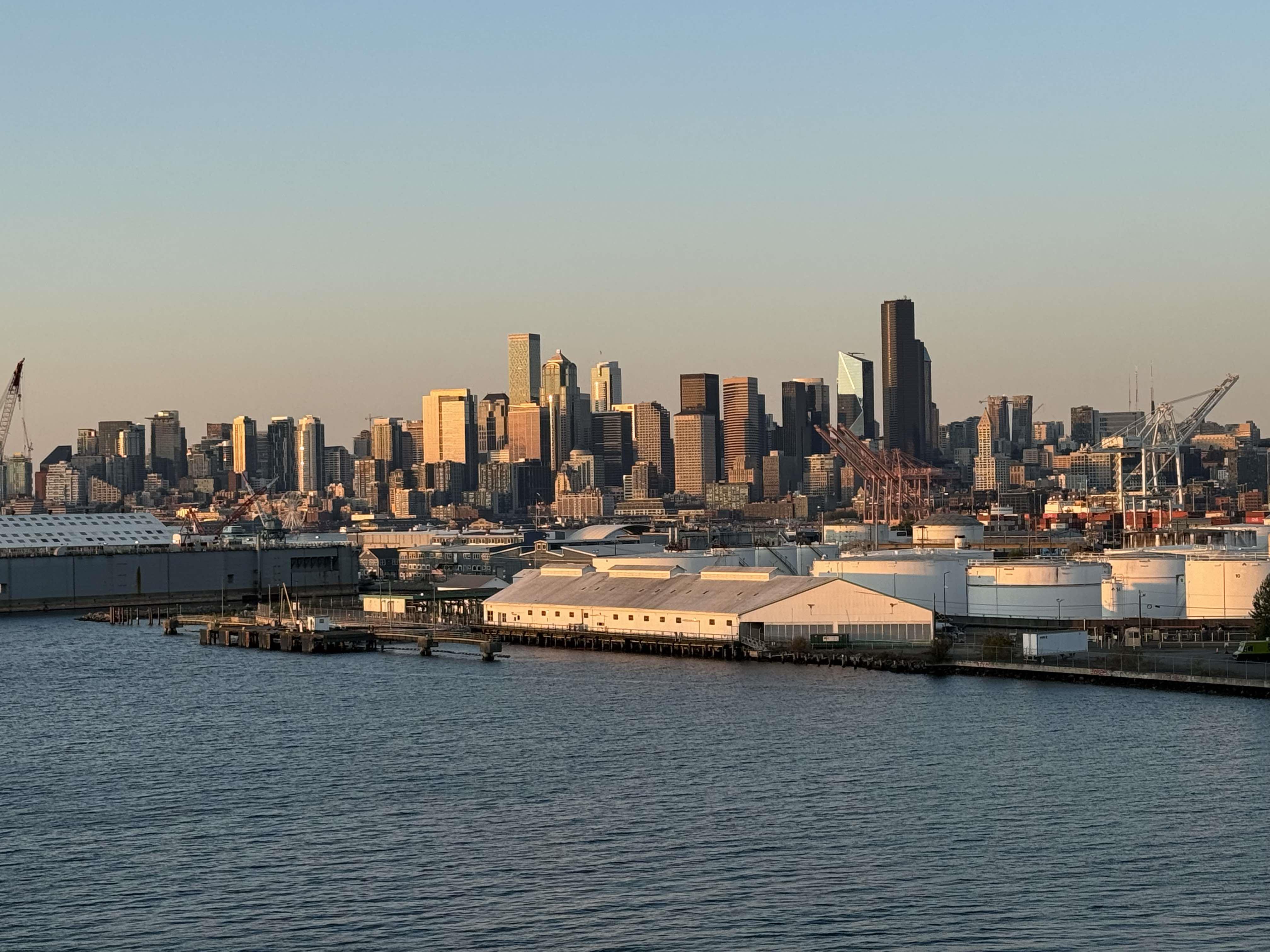 The city skyline glows warmly as the sun sets over the calm waterfront and industry.
