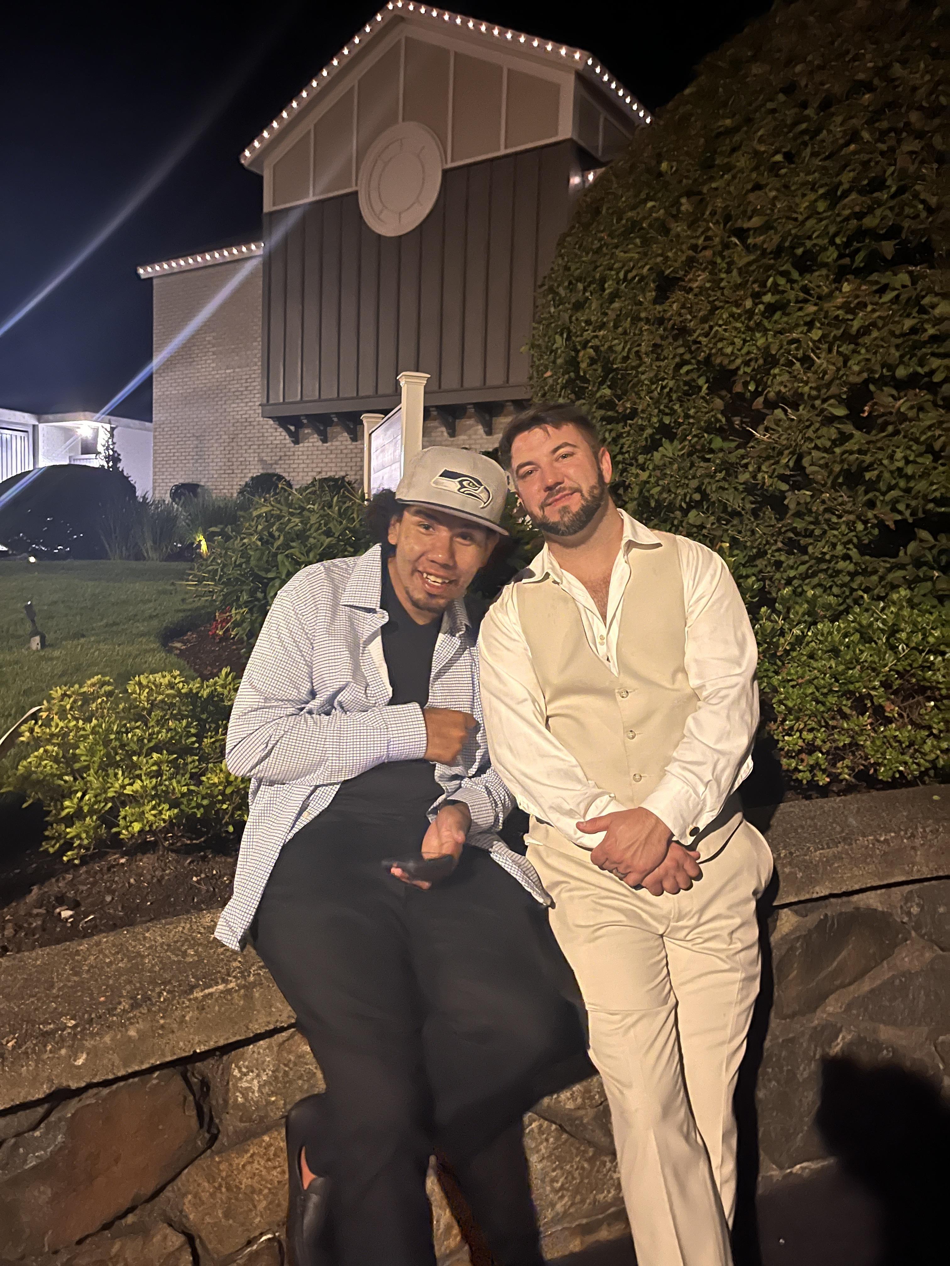 Two friends pose happily outside a well-lit venue at night, surrounded by plants and atmosphere.