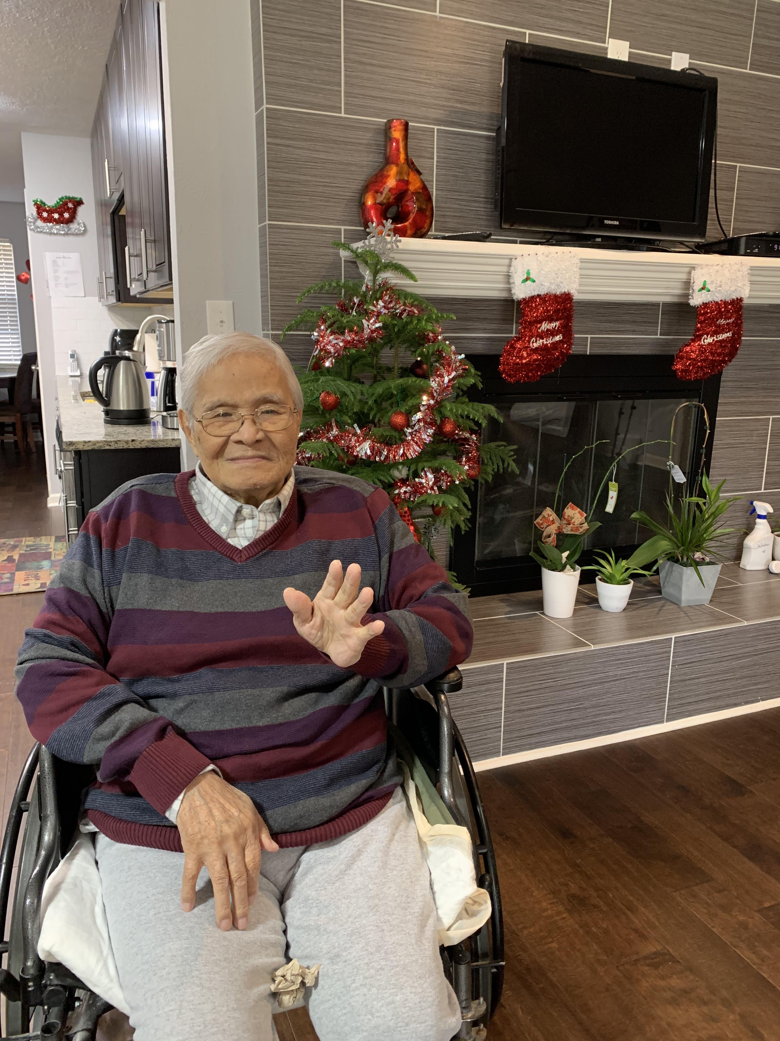 An elderly man in a wheelchair greets with a smile amid festive decorations in a warm living space.