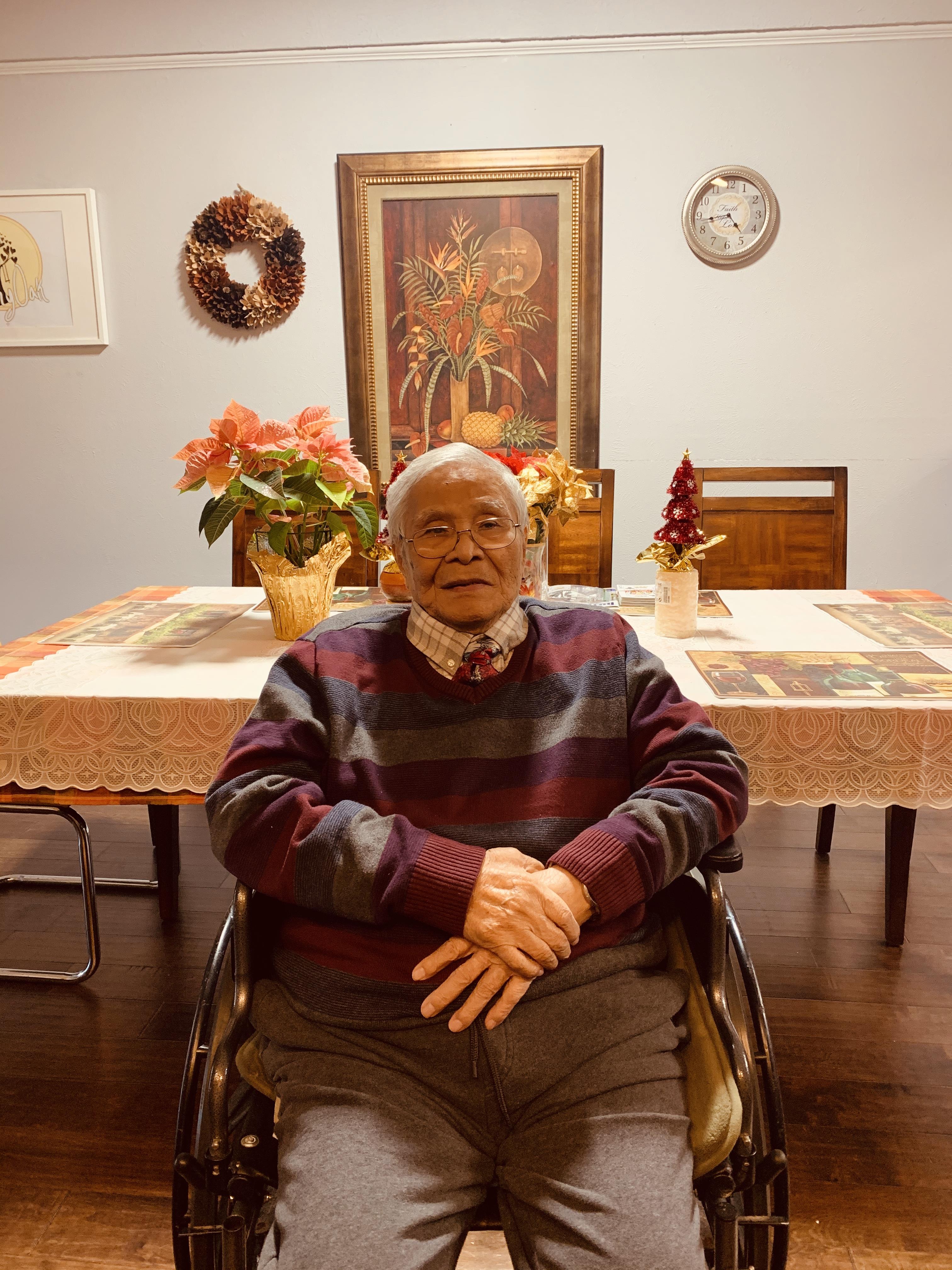 An elderly man in a wheelchair relaxes in a warm dining room filled with plants and festive decor.