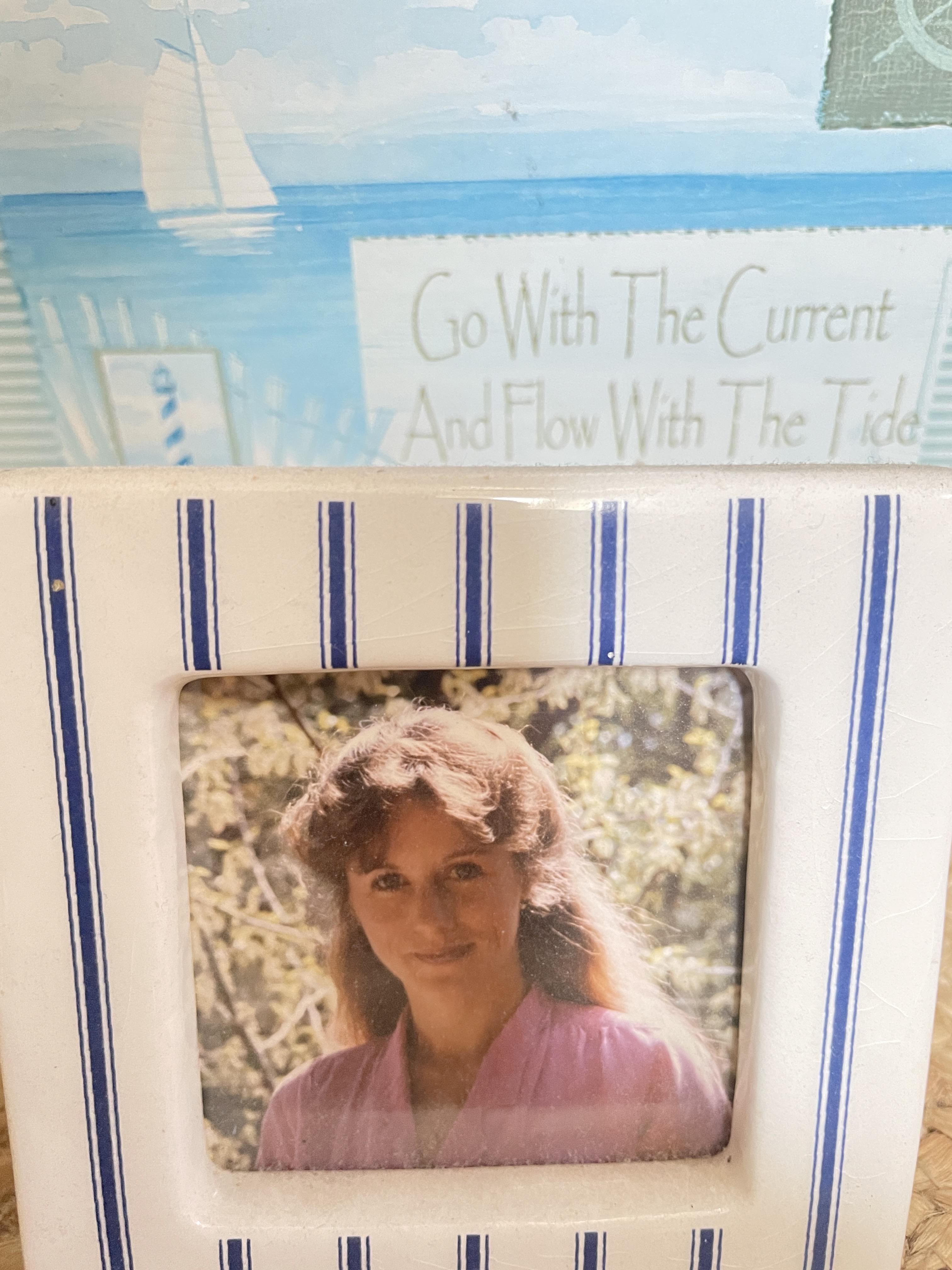A woman stands smiling in a pink top, surrounded by foliage and a calm water view.