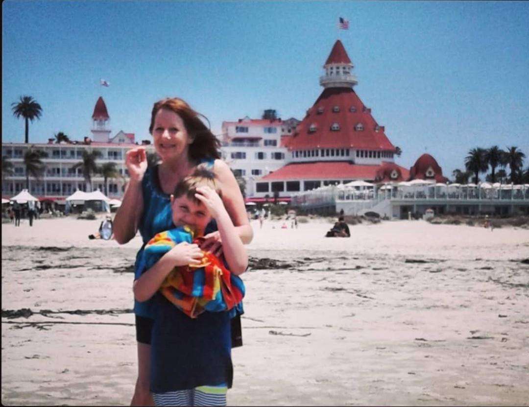 A mother and her son are standing on the sandy beach, smiling brightly at the sunny atmosphere.
