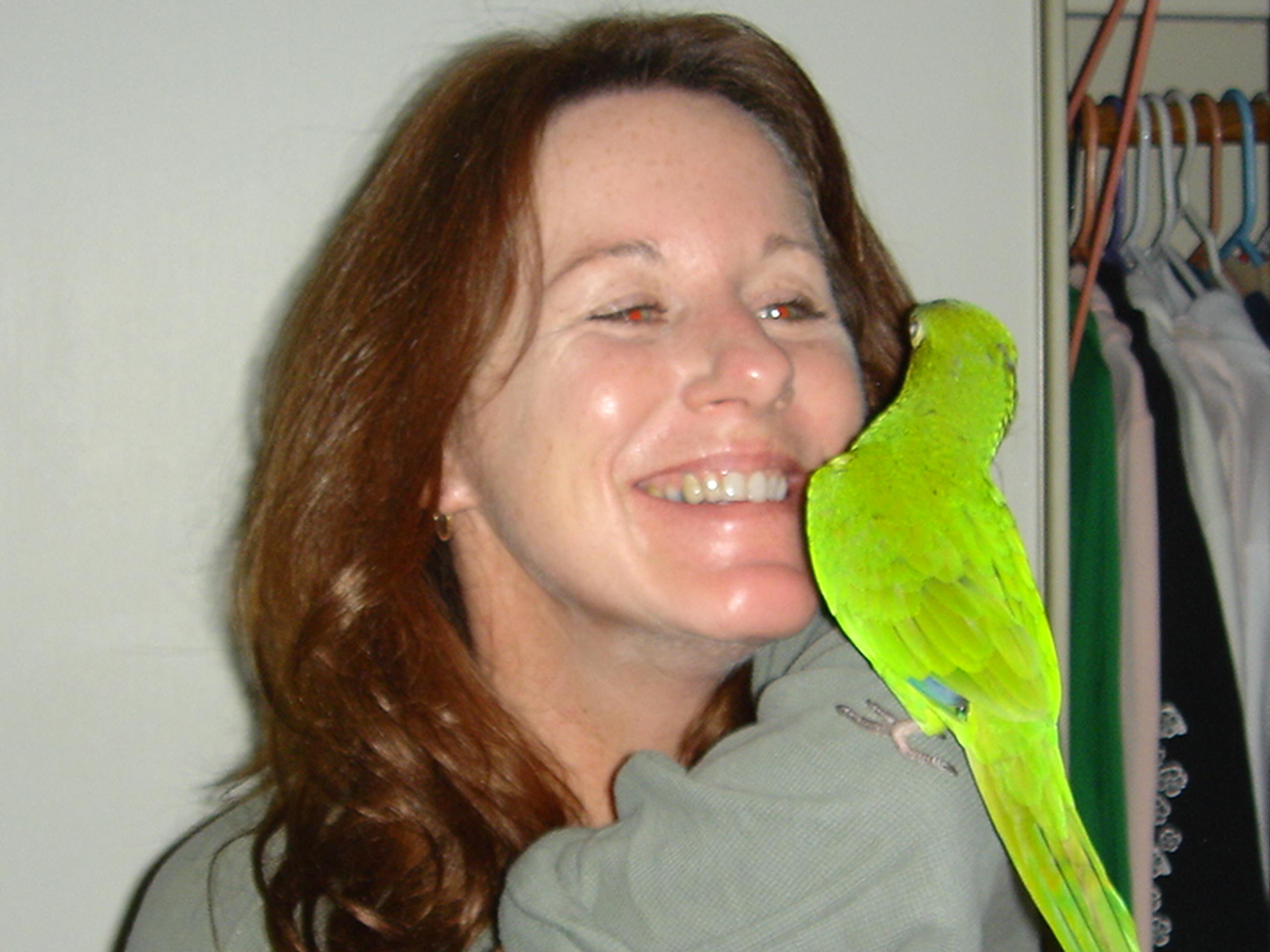 Woman smiles as a vibrant green parrot perches playfully on her shoulder indoors.