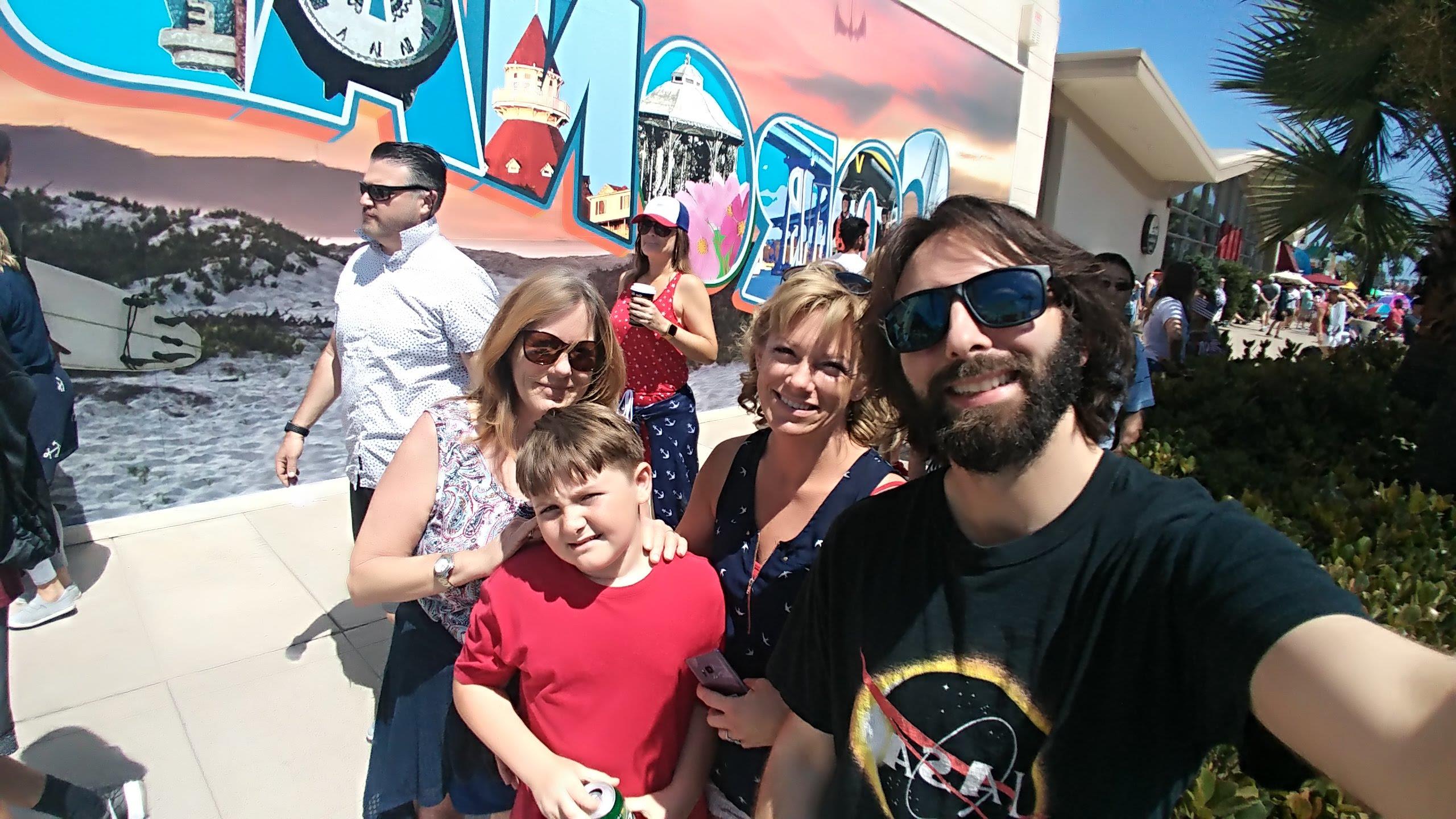 Group of families smiling and posing for a picture near a vibrant mural on a sunny day at the beach.