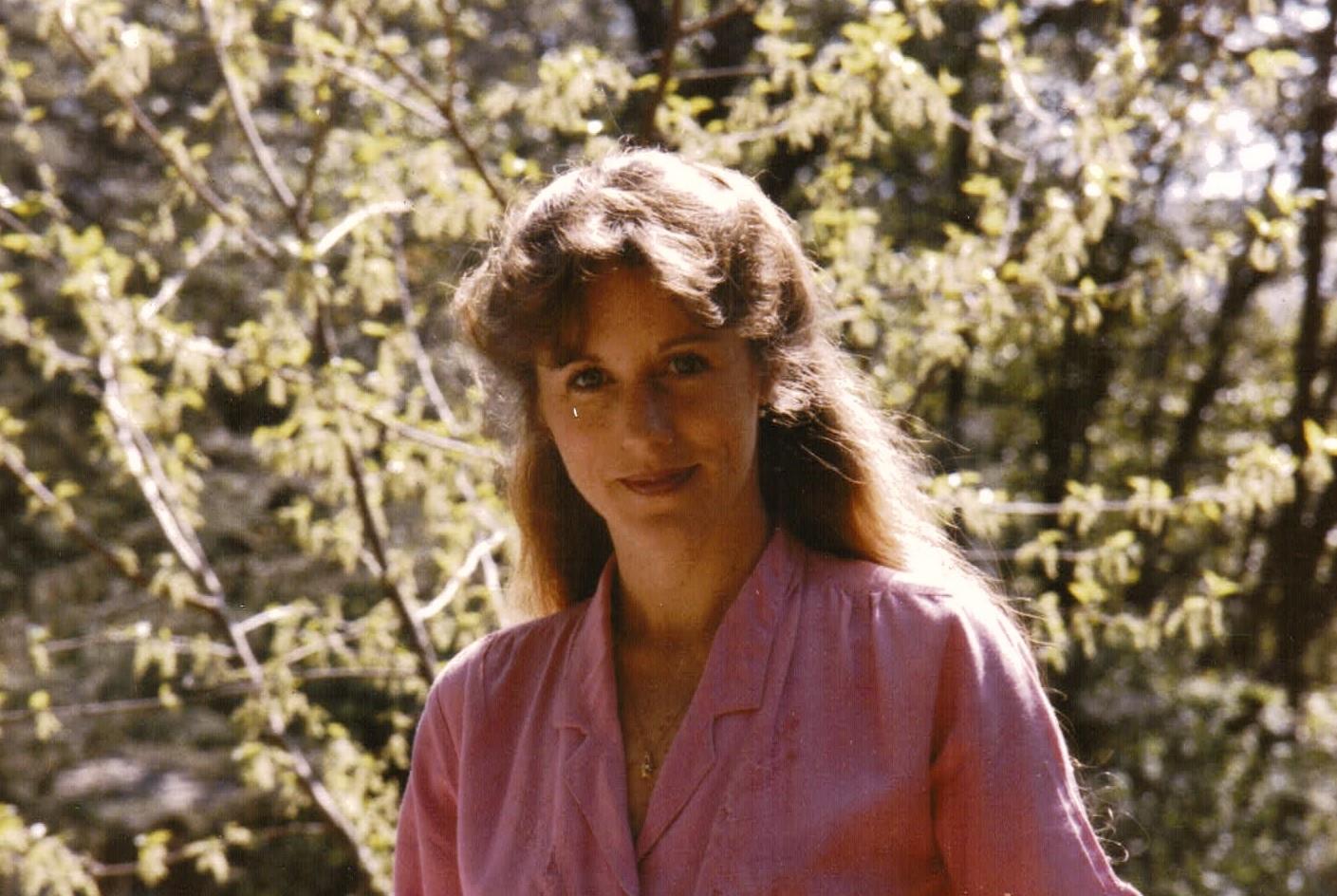 A woman poses outside in a pink blouse surrounded by greenery and sunlight in early spring.